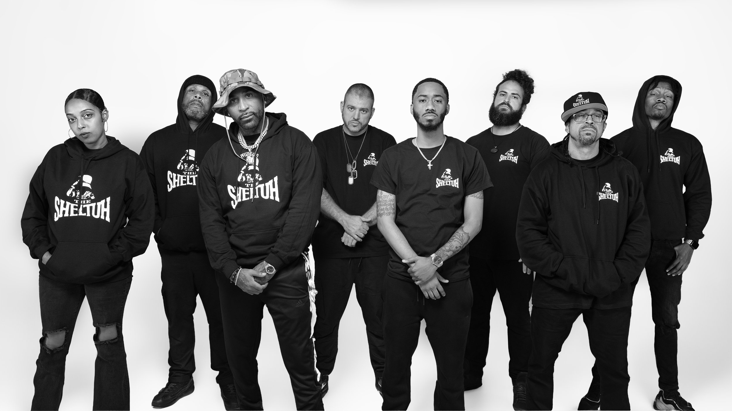 Group of nine diverse people standing against a plain white background, wearing black hoodies and T-shirts with the logo 'The Sheluth'. BoldVision Studios