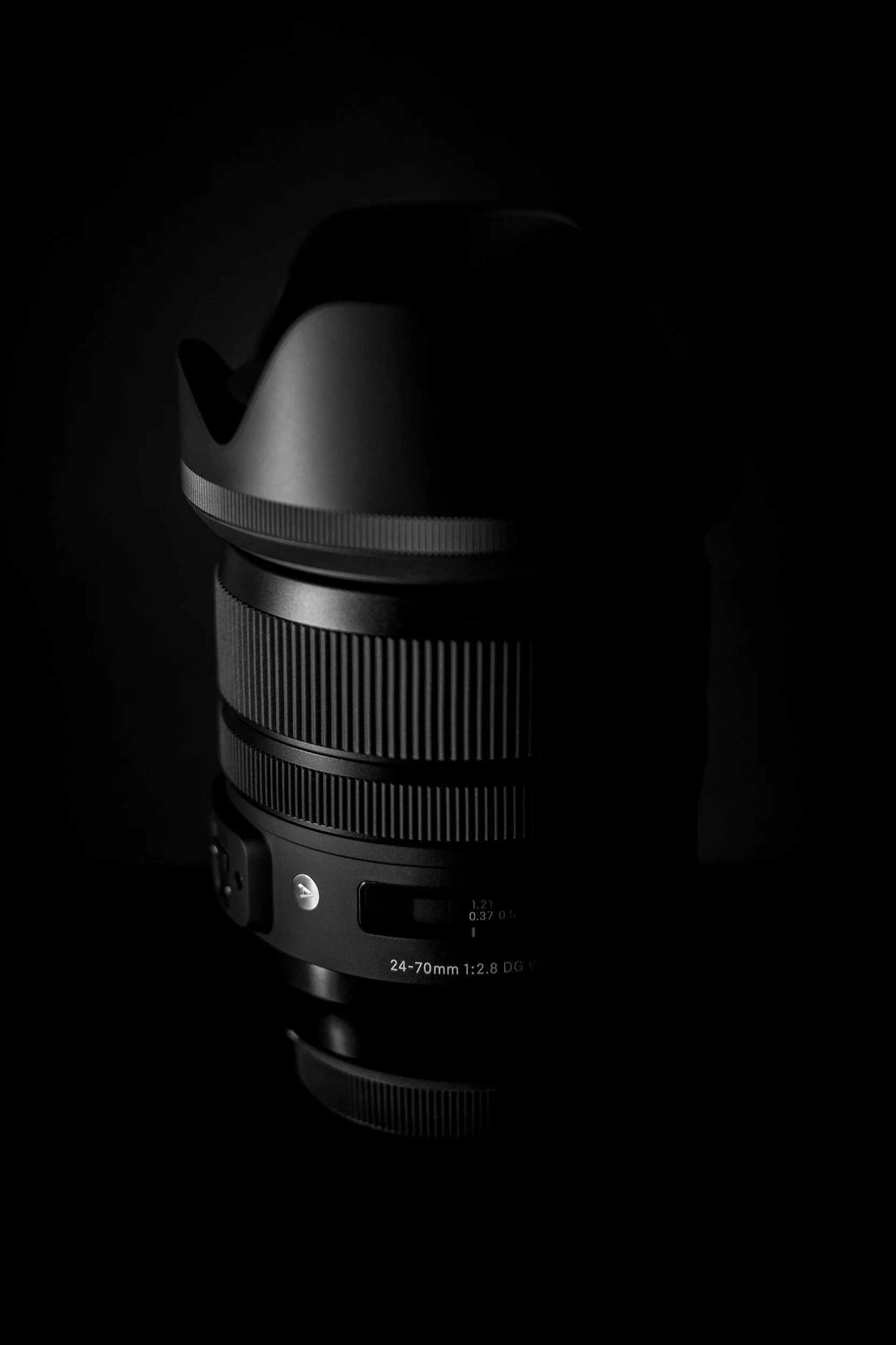 Black camera lens with a lens hood, against a dark background, illuminated to highlight its details.