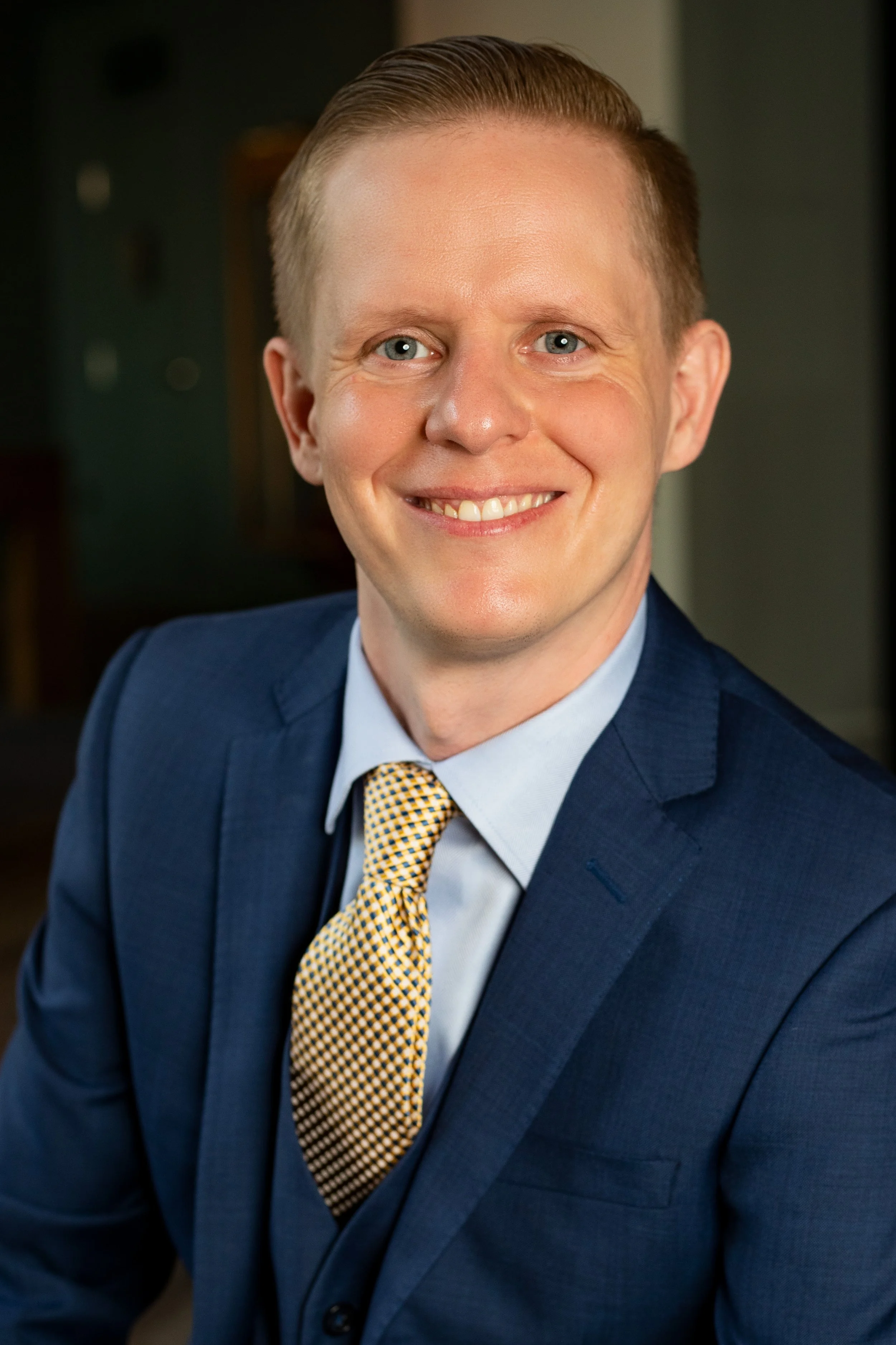 Headshot of a man in a blue suit, light blue shirt, and patterned yellow tie, smiling against a blurred indoor background. BoldVision Studios