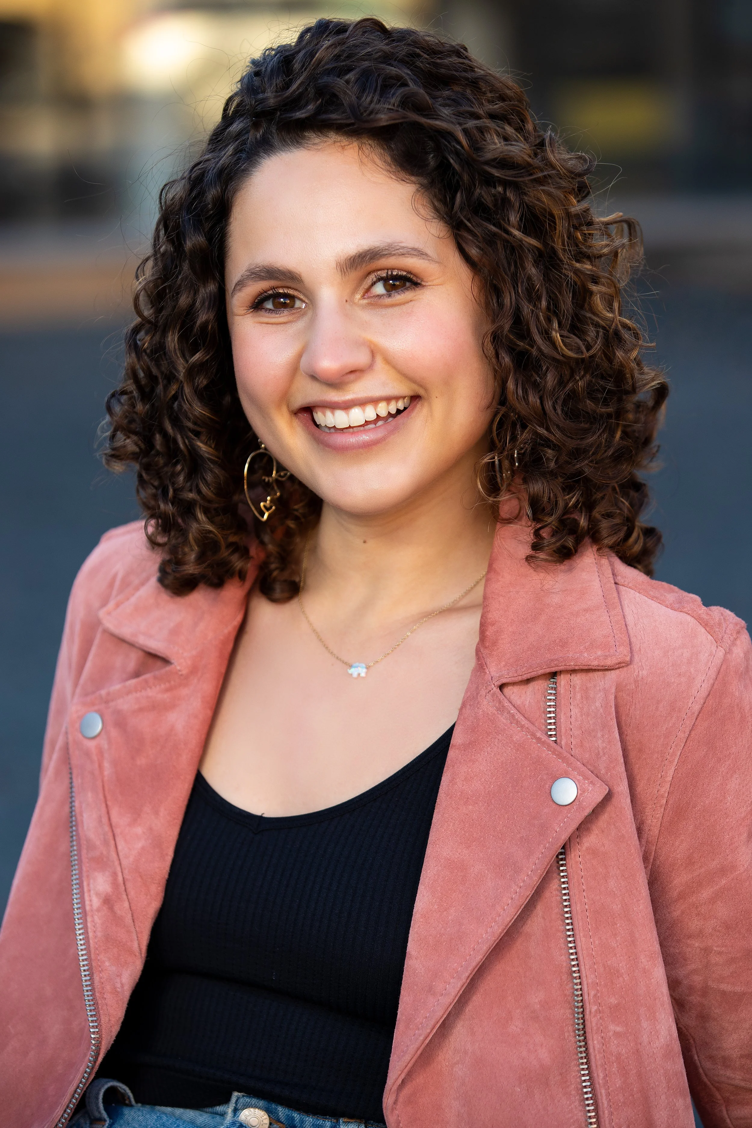 A young woman with curly brown hair smiling, wearing a pink jacket, black top, and jewelry, outdoors during daytime. BoldVision Studios
