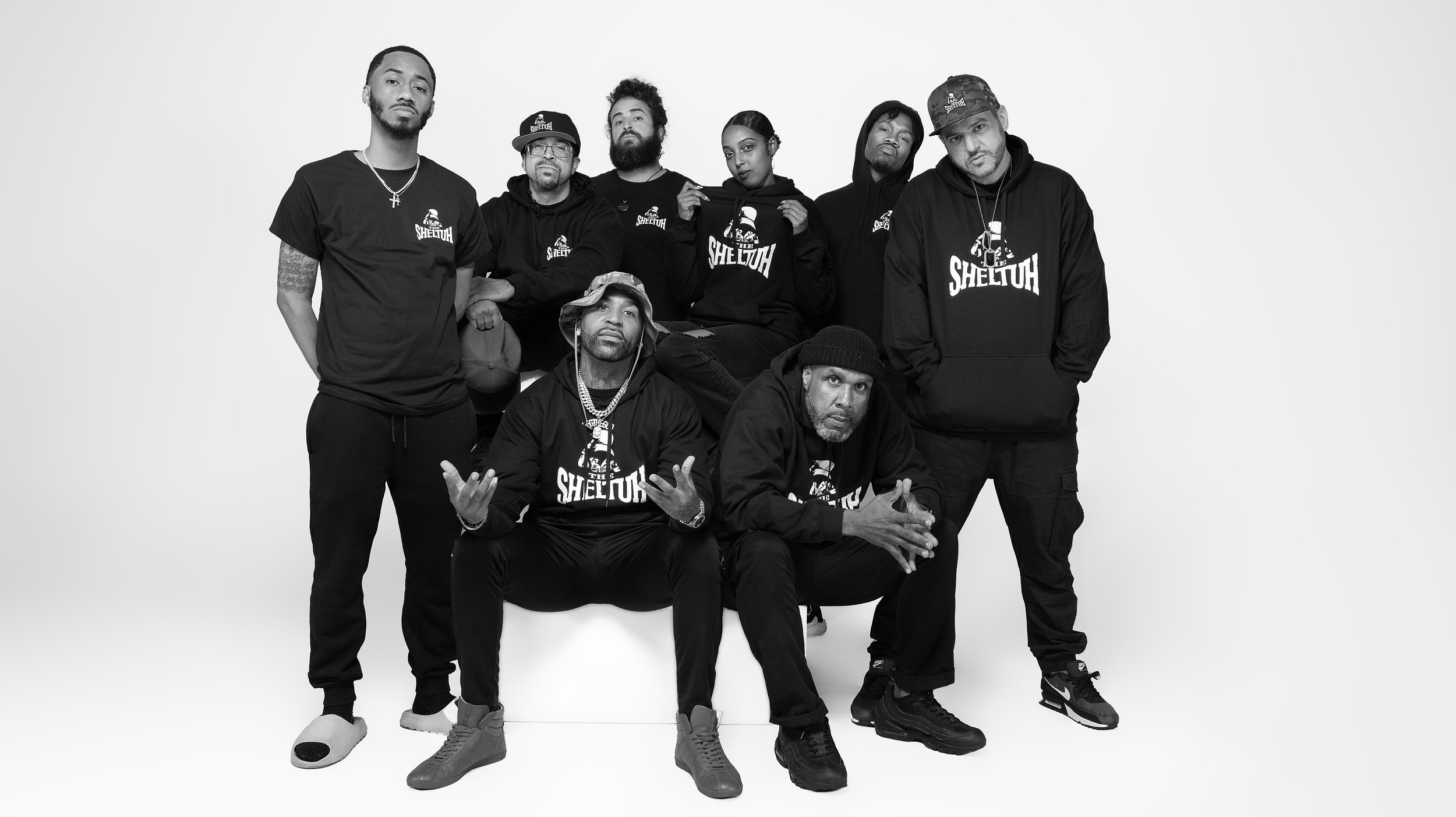 Black and white photo of nine diverse individuals wearing matching hoodies and t-shirts with the word 'SHELUTH' printed on them, posed in a studio with a plain white background. BoldVision Studios