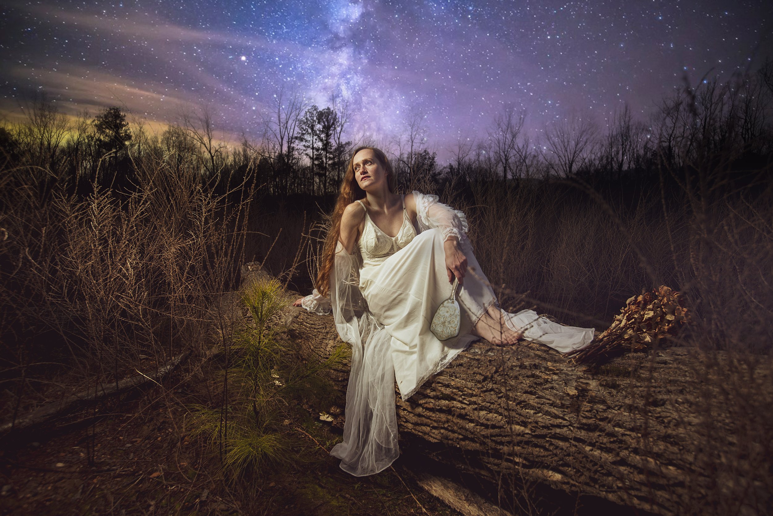 A woman in a white dress with long hair sitting on a fallen tree trunk in a forest clearing at night, with a starry sky and the Milky Way galaxy visible in the background. BoldVision Studios