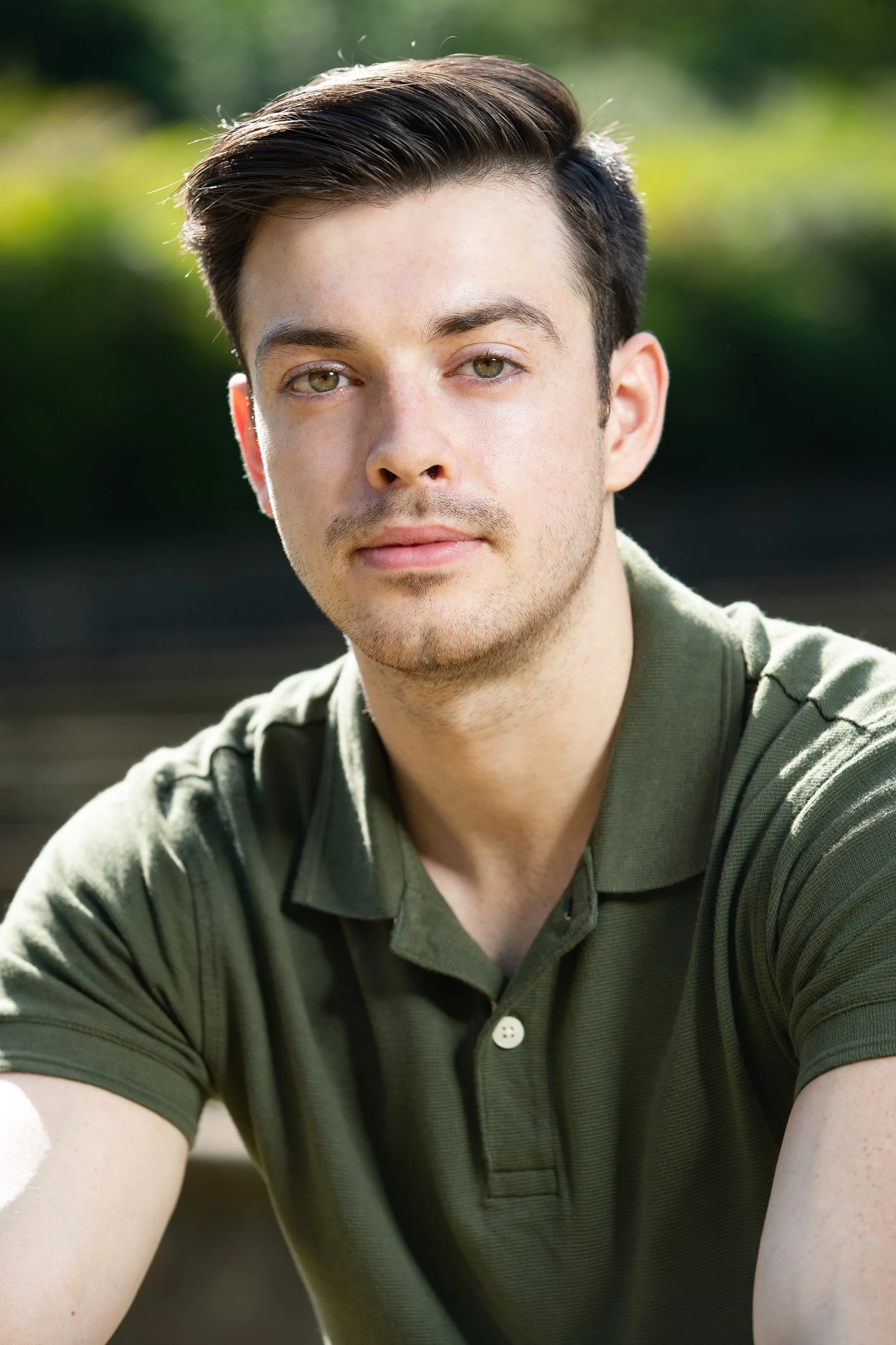 Close-up of a young man with light skin, short brown hair, and hazel eyes, wearing a green polo shirt outdoors on a sunny day with a blurred green background.