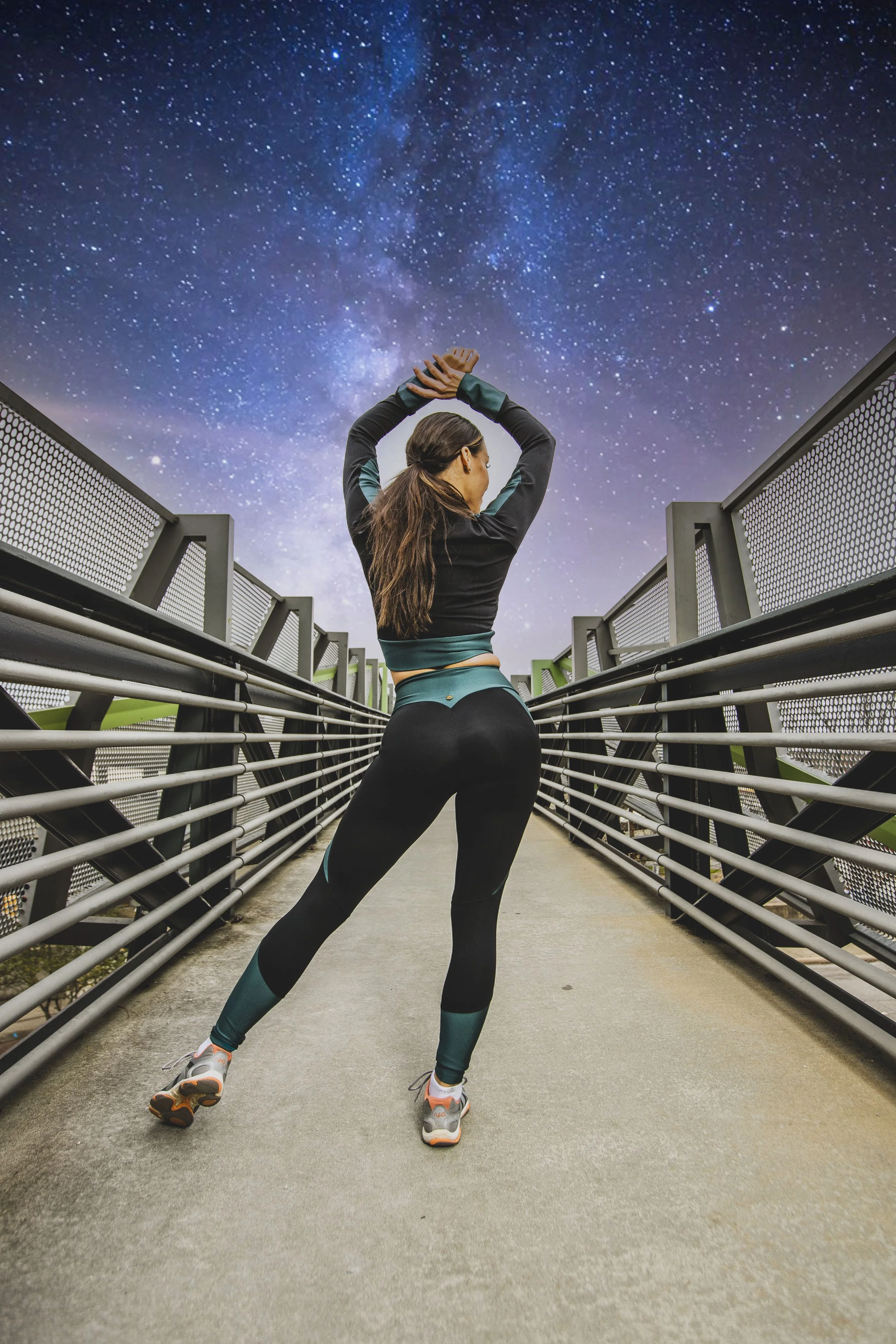 A woman standing on a bridge at night with her back to the camera, looking up at a starry sky and the Milky Way galaxy above. BoldVision Studios