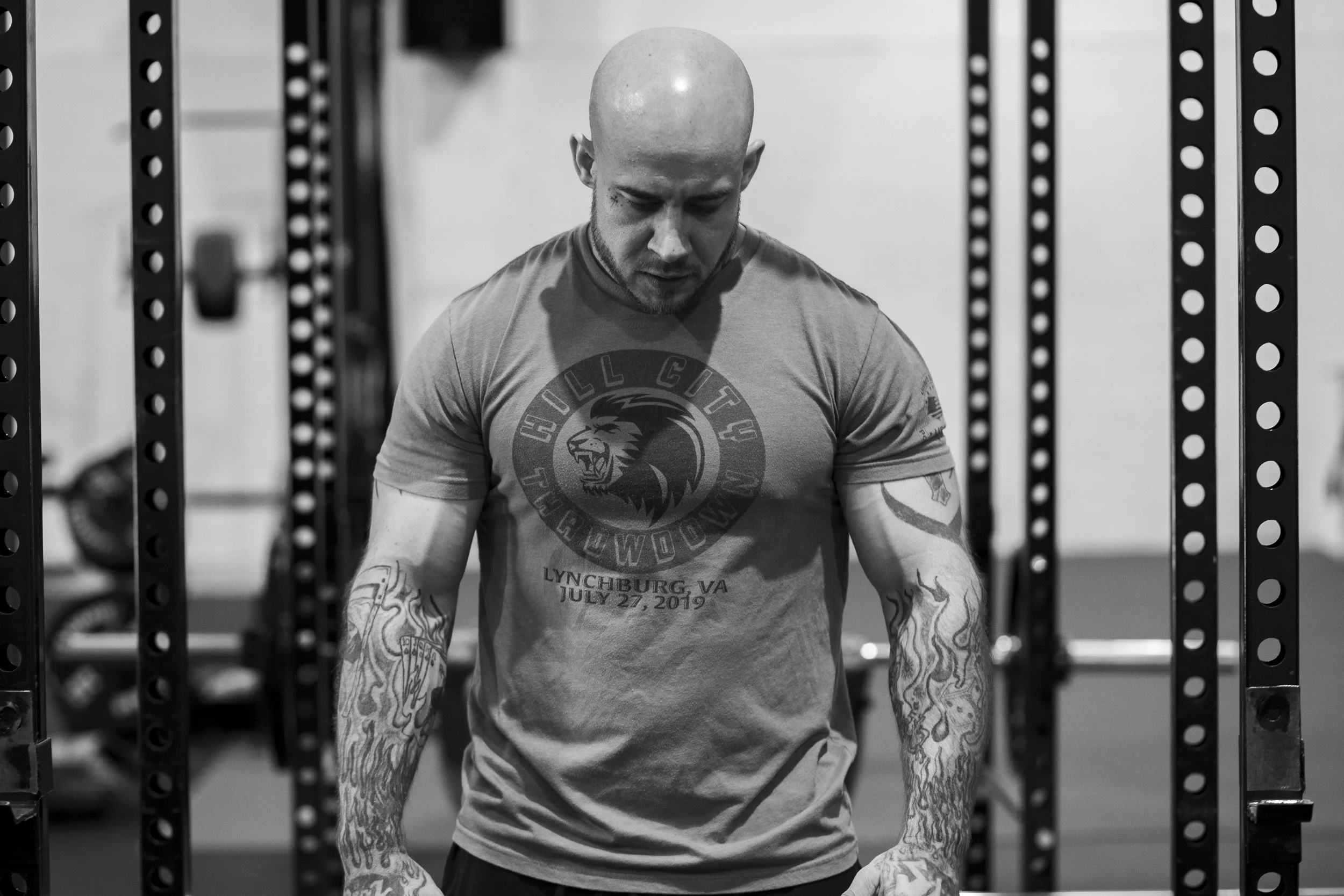 A muscular, bald man with tattoos on both arms is looking down while standing between weightlifting racks at a gym. BoldVision Studios