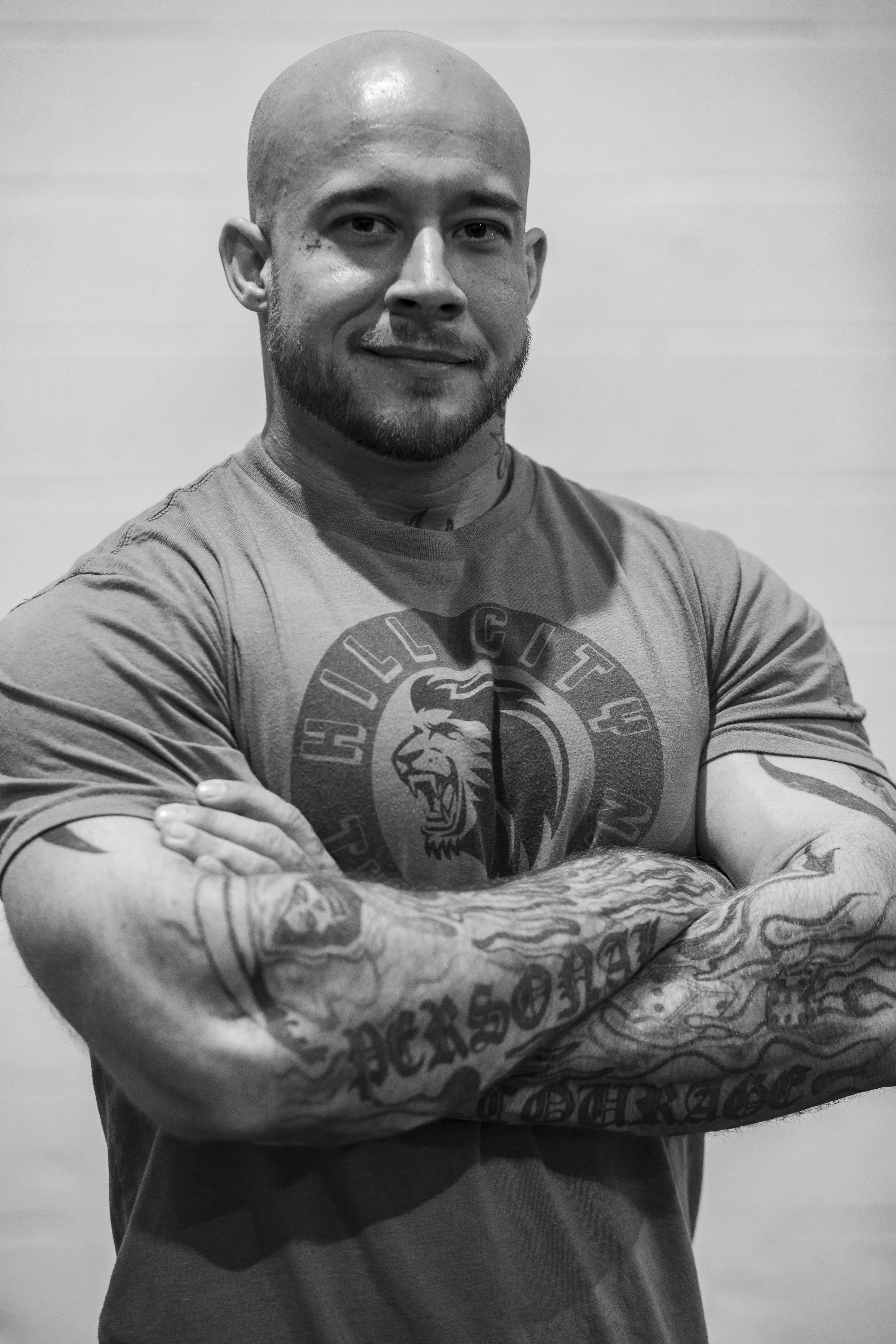 A muscular man with tattoos on his arms and neck, smiling and standing with his arms crossed. He is wearing a t-shirt with a lion's head logo and the word 'Bulldog' visible. BoldVision Studios