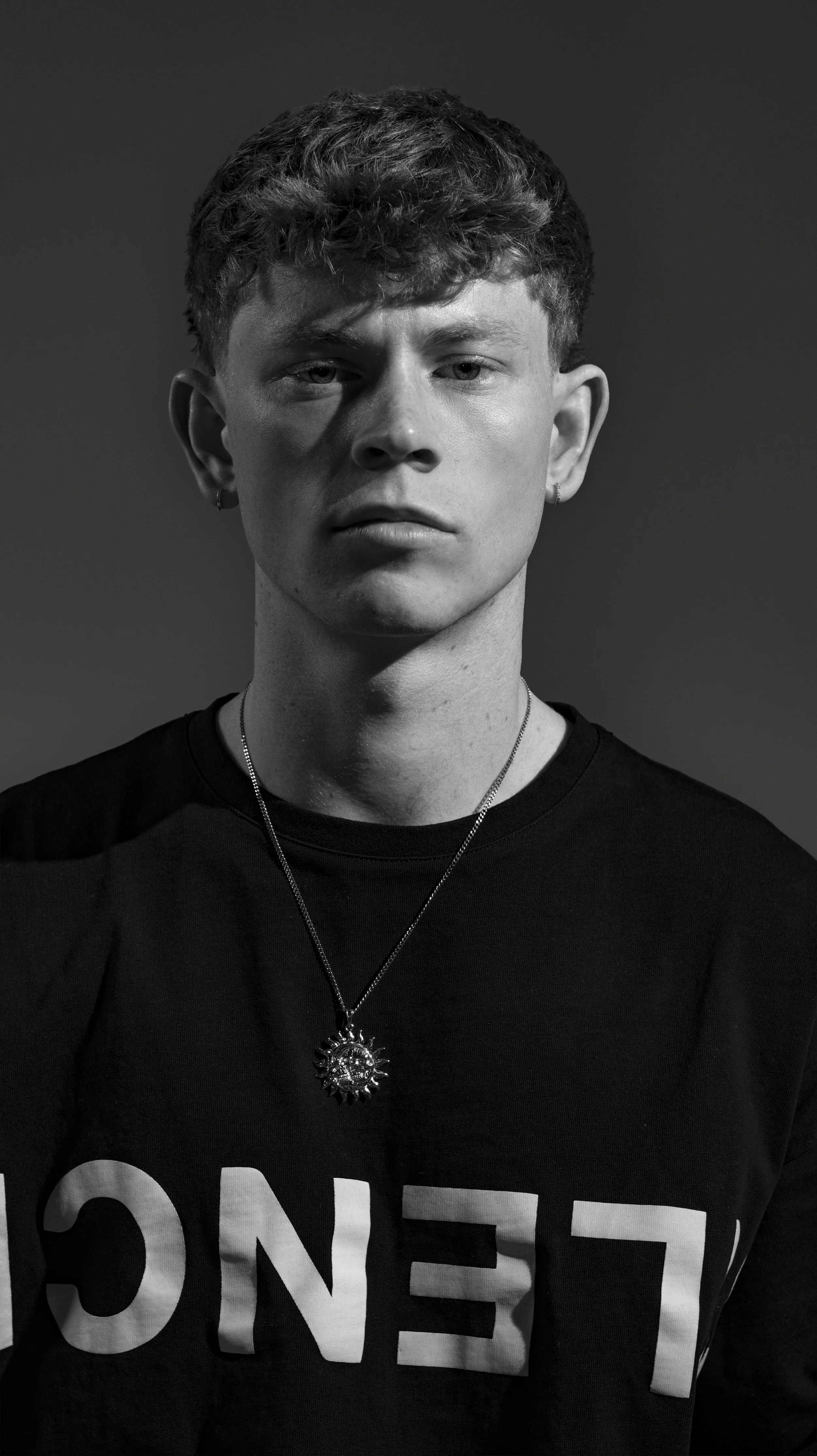 Black and white portrait of a young man with short curly hair, wearing a black shirt with white text and a necklace with a sun-shaped pendant. BoldVision Studios