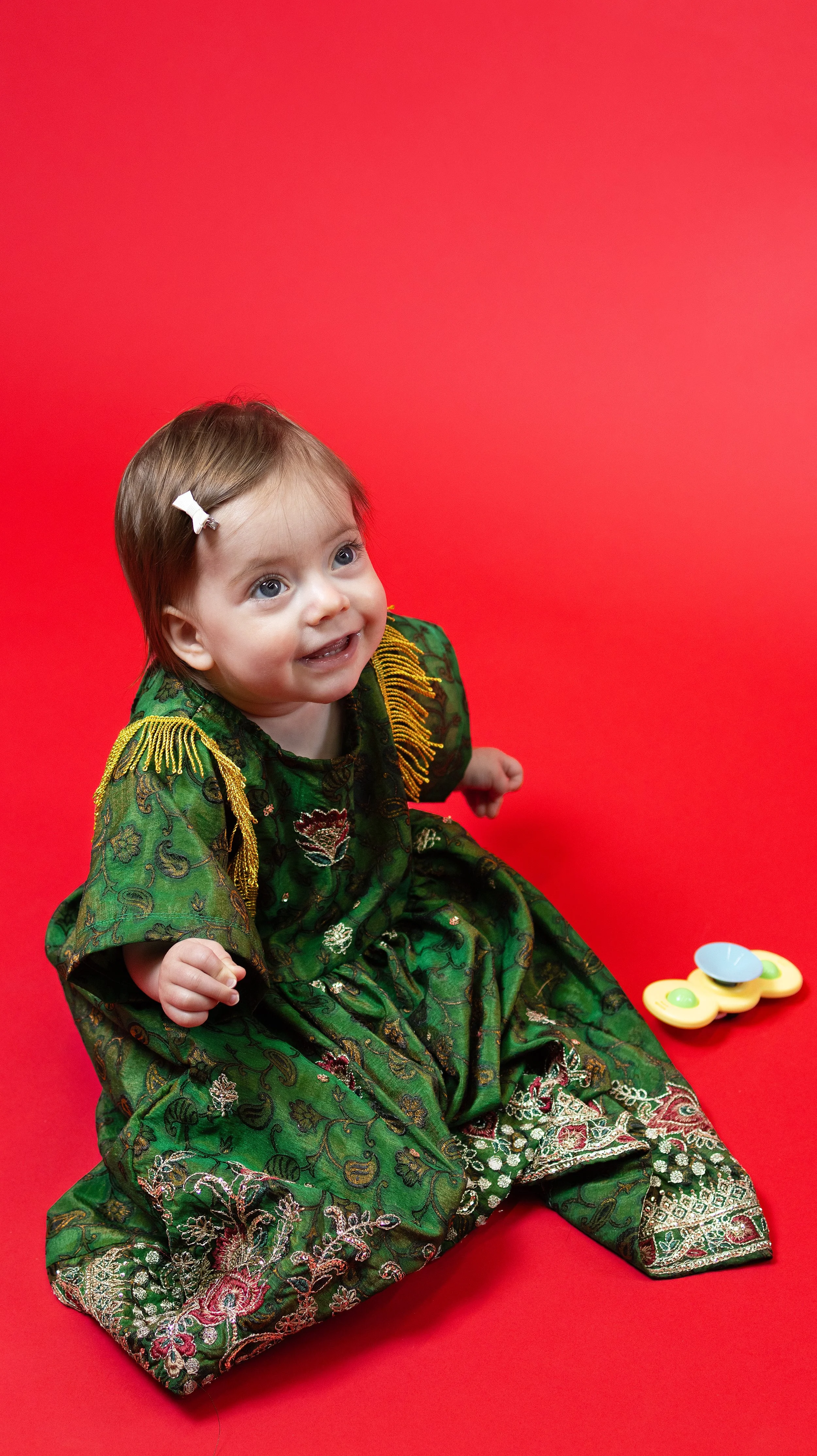 A young girl with brown hair, wearing a green traditional dress with intricate embroidery and gold fringe, sitting on a red background next to a yellow fidget spinner toy with a blue scoop. BoldVision Studios