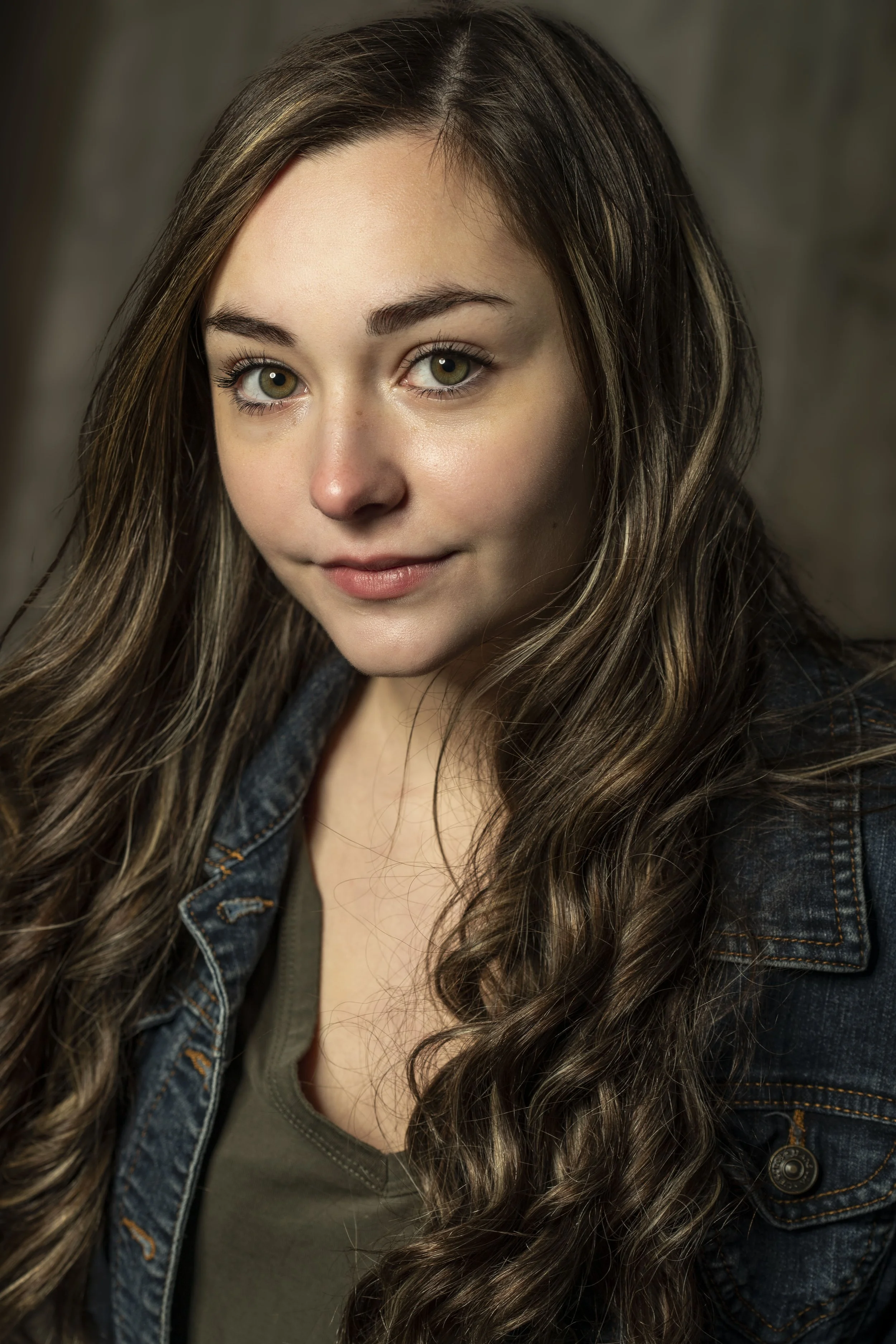 Close-up of a young woman with long, wavy brown hair and hazel eyes, wearing a denim jacket and an olive green top, looking at the camera with a slight smile. BoldVision Studios