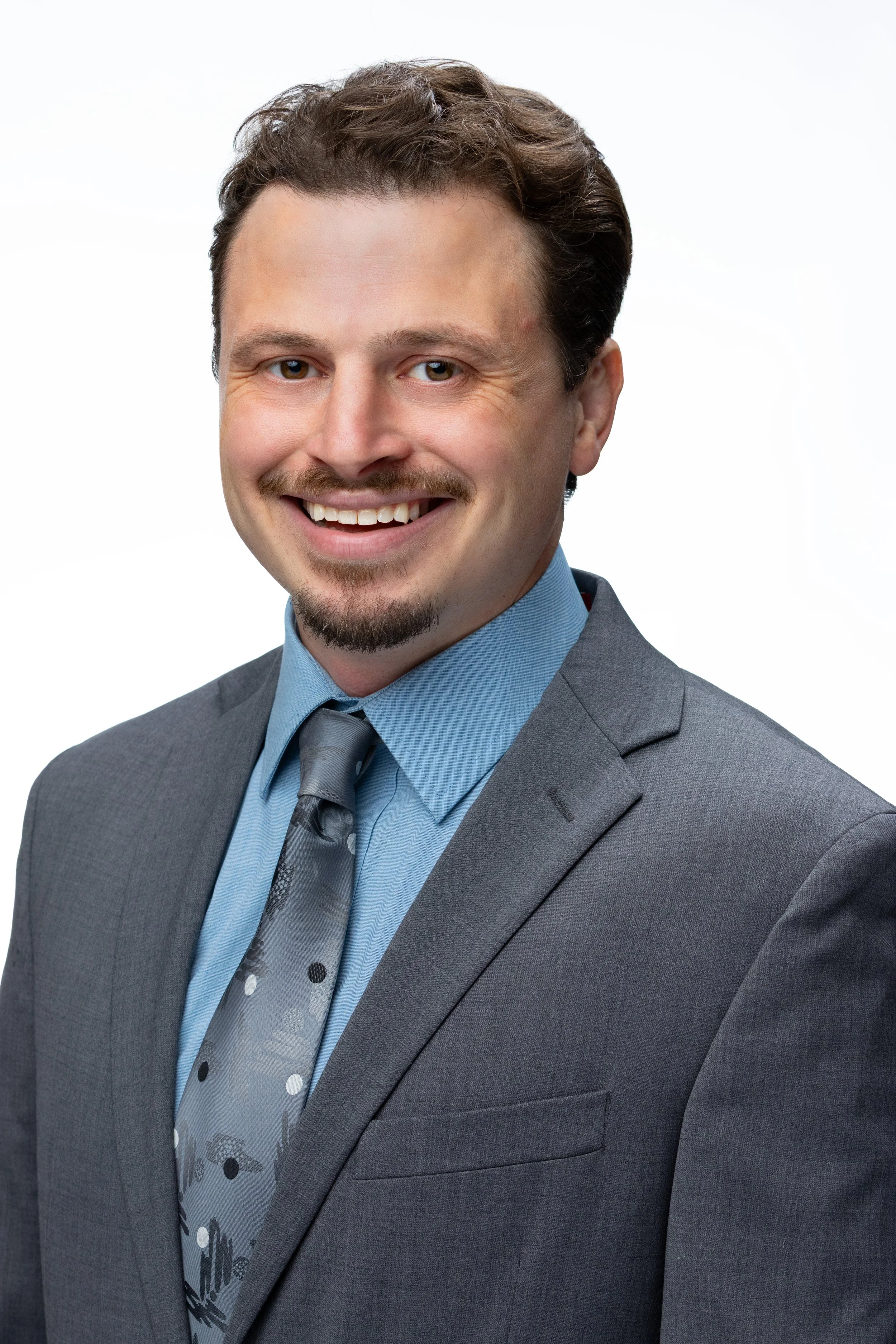 A portrait of a man with light skin, brown curly hair, and a mustache, smiling in a professional gray suit, light blue shirt, and patterned tie against a plain white background.