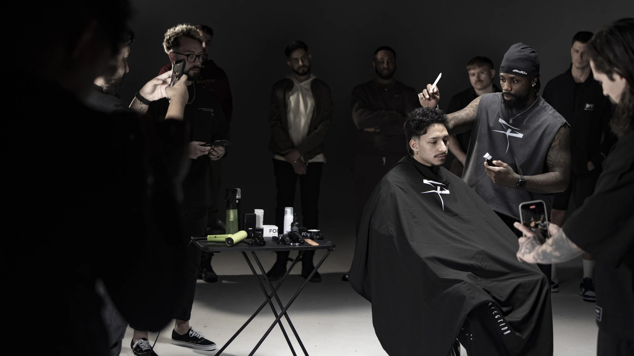 A group of people surrounding a man getting a haircut in a dark studio, with hairstyling tools on a small table in the foreground. BoldVision Studios