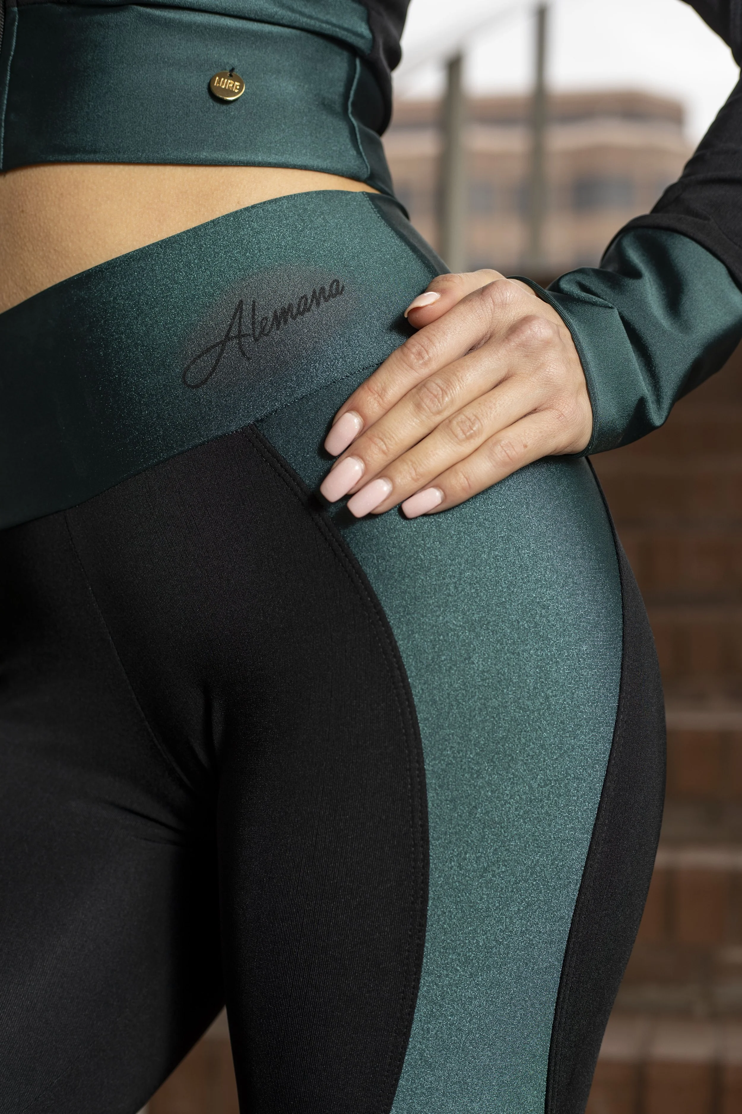 Close-up of a woman wearing athletic leggings and a matching long-sleeve top, with her hand resting on her hip. The leggings have a signature logo, and the waistband features a small gold pin labeled 'Lure.' BoldVision Studios