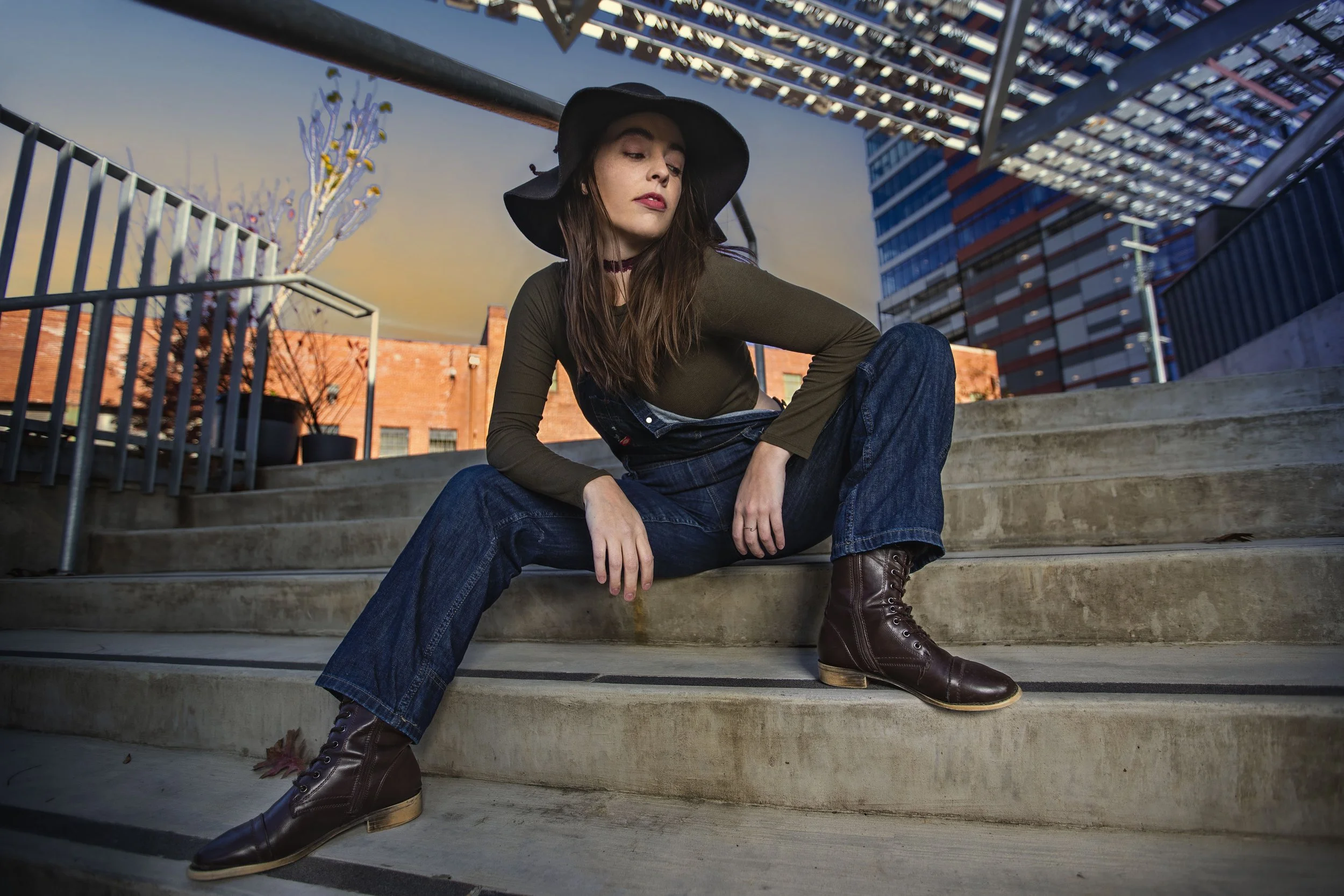 A young woman sitting on outdoor concrete steps in an urban setting, wearing a black hat, green long-sleeve shirt, blue jeans, and brown lace-up boots, with modern buildings and a sunset sky in the background. BoldVision Studios