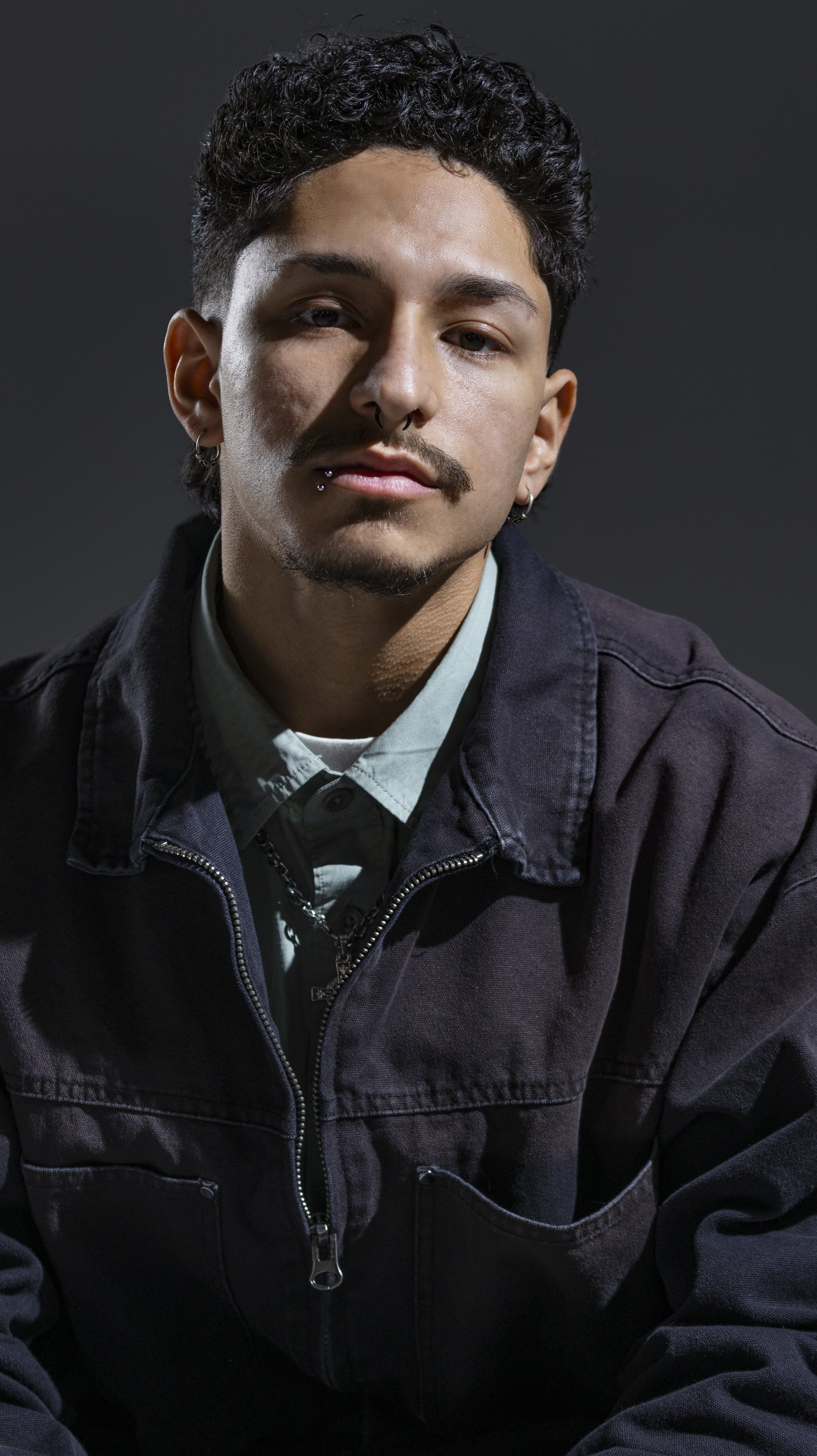 A young man with short curly dark hair, a mustache, and piercings on his lower lips and ears, wearing a black jacket and a mint green shirt, looking at the camera against a dark gray background. BoldVision Studios
