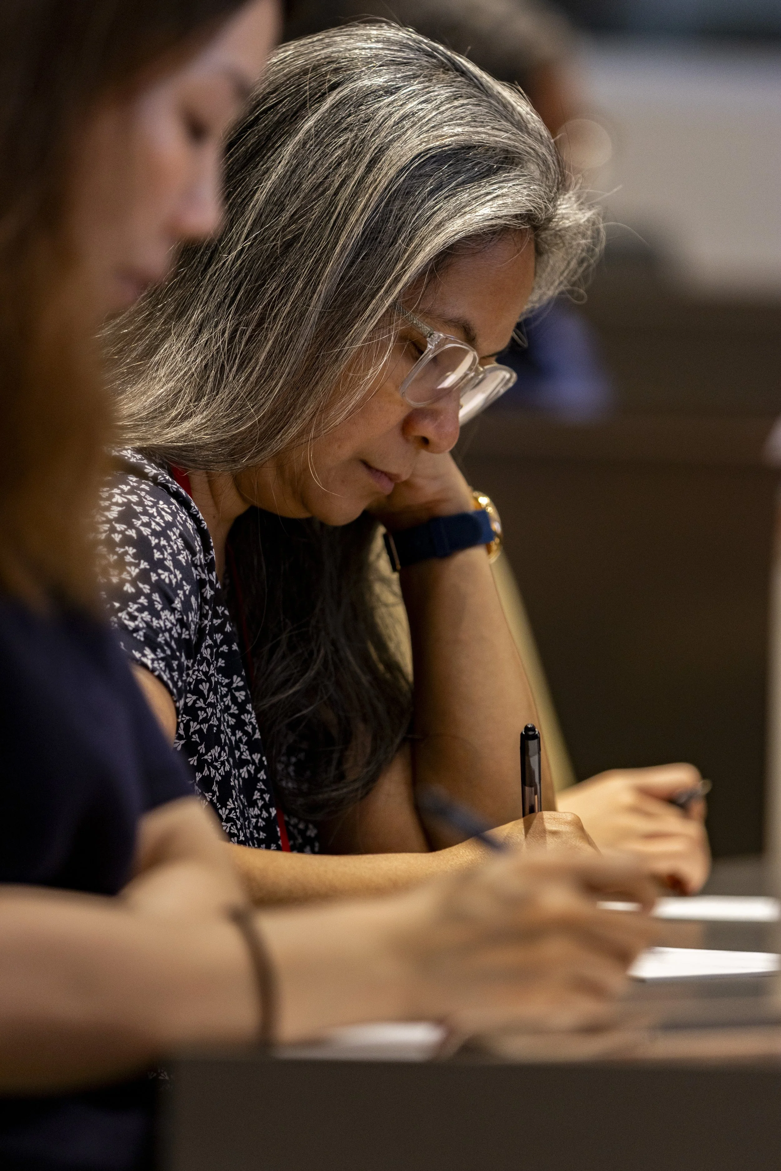 Close-up of a woman with long gray hair, wearing glasses, sitting at a table and taking notes with a pen during a meeting or conference.