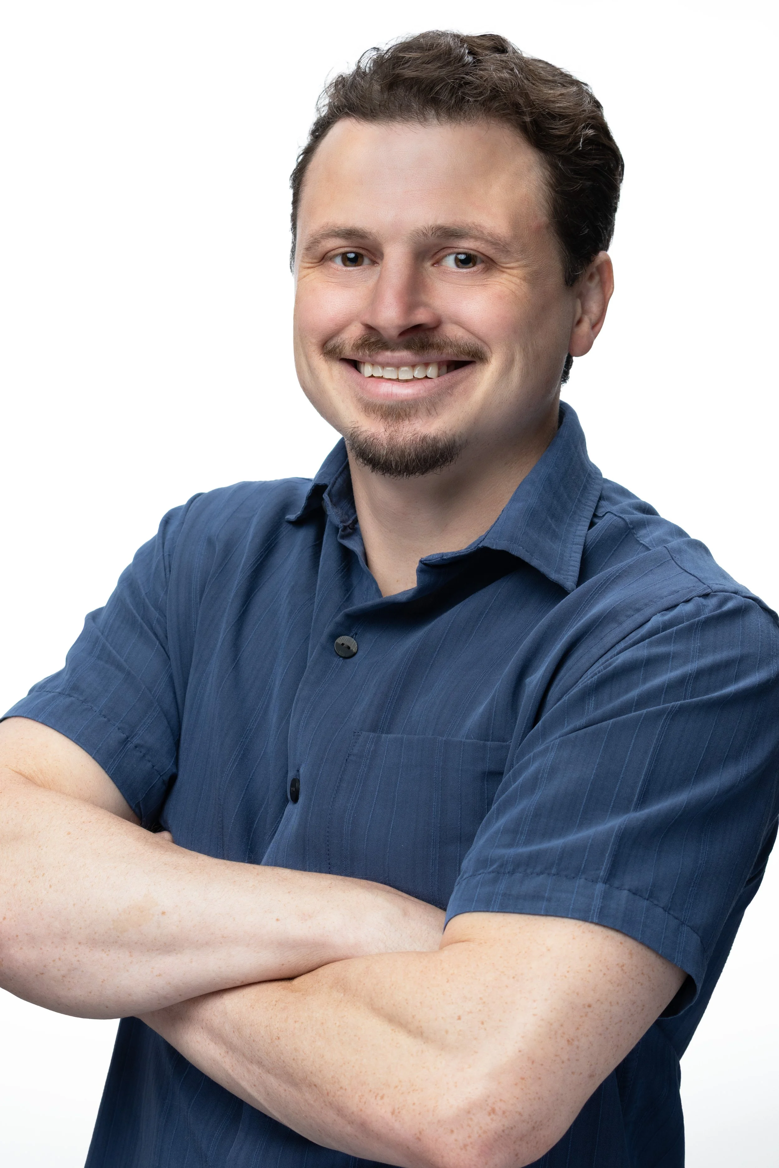 A man with brown hair, a mustache, and goatee, wearing a blue collared shirt, smiling, with arms crossed against a white background.