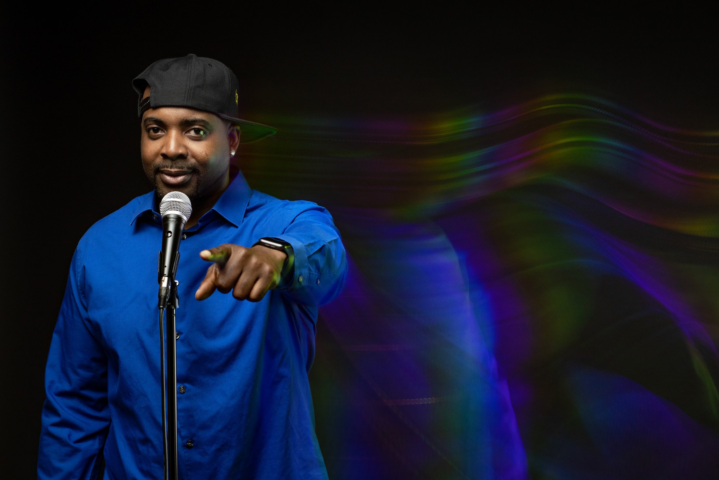 A man wearing a black baseball cap and a blue shirt is pointing towards the camera while standing in front of a microphone. The background is black with colorful, flowing light streaks. BoldVision Studios