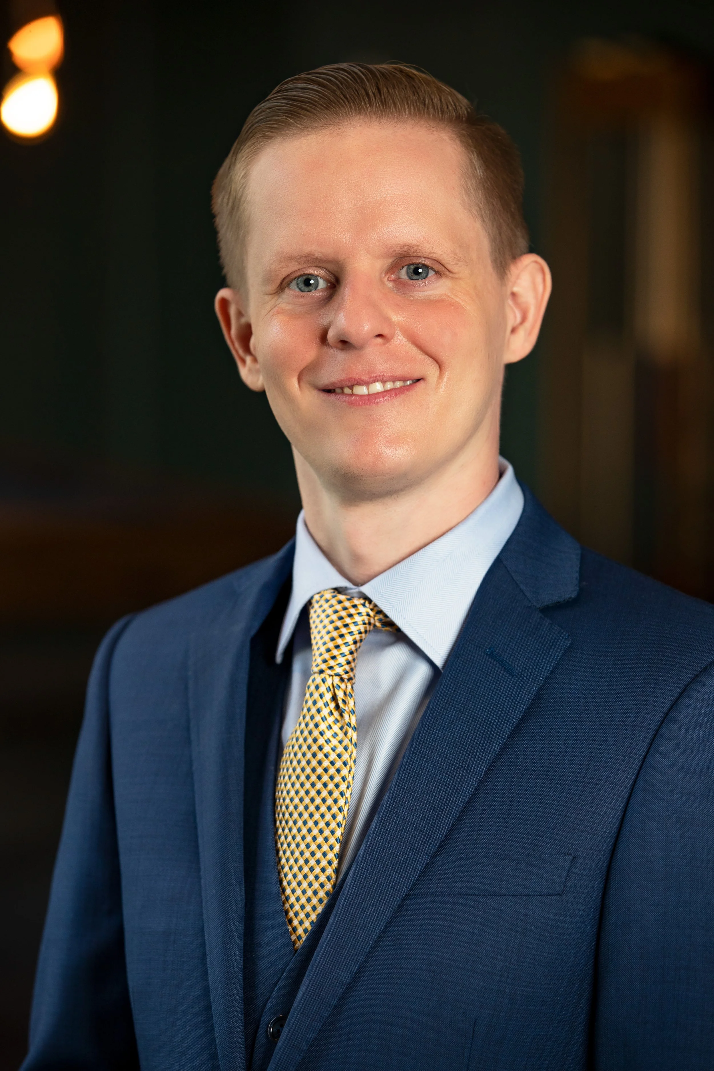 A professional headshot of a man in a blue suit, light blue dress shirt, and a yellow checkered tie, smiling against a dark background with warm lighting. BoldVision Studios