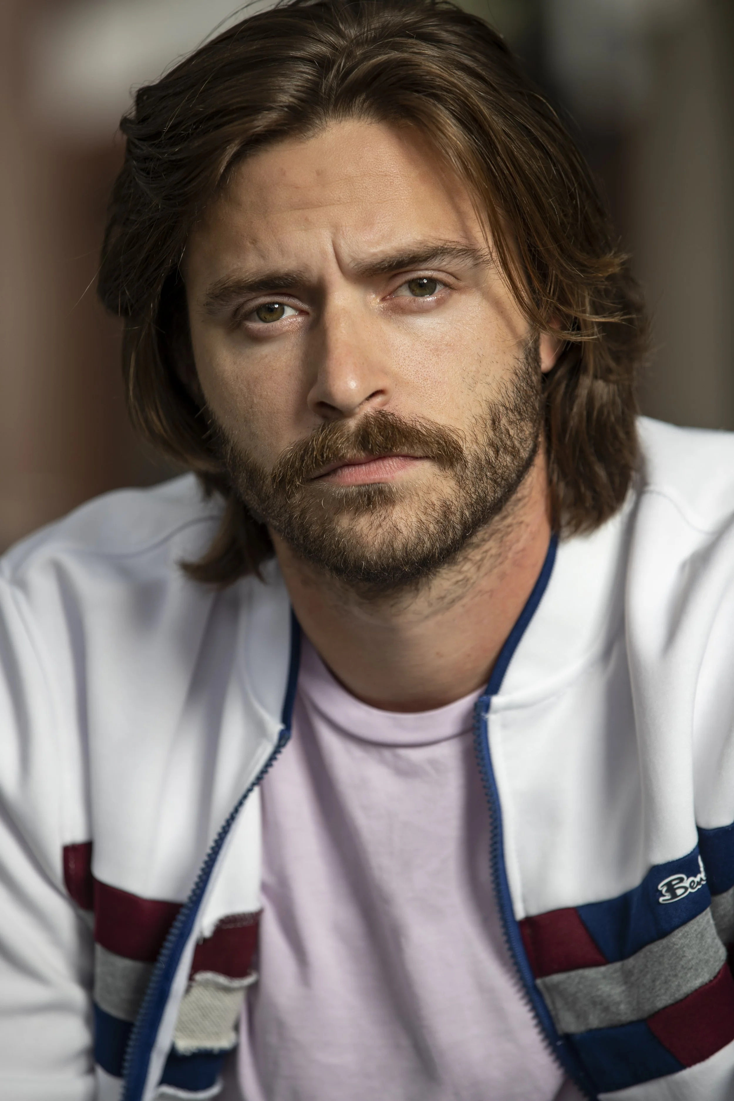 Close-up of a man with brown hair, beard, and mustache, wearing a white and multicolored zip-up jacket and a pink shirt underneath.