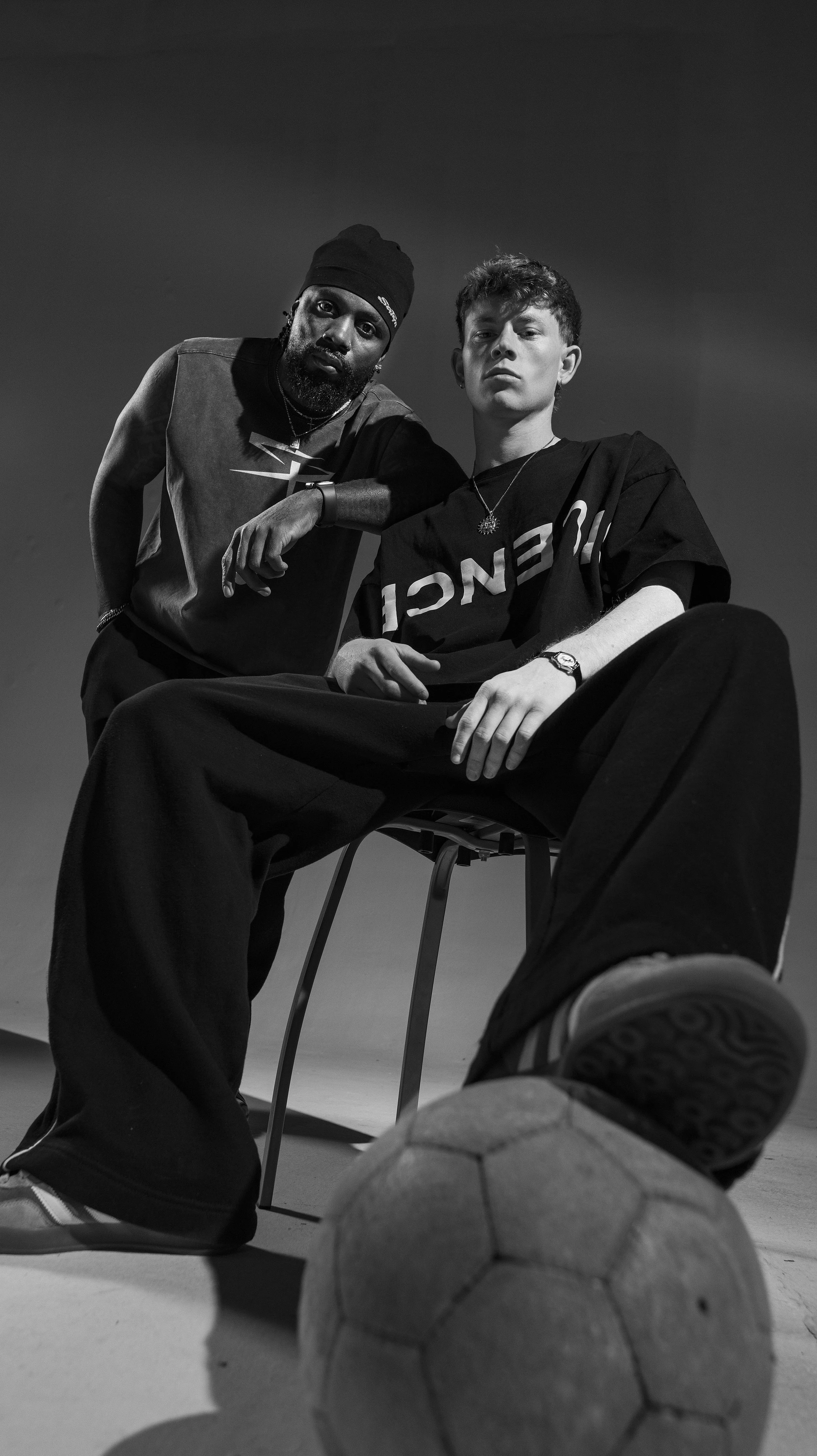 Black and white photo of two young men, one sitting on a chair with a soccer ball near his foot, and the other leaning on the chair's back, in a studio setting. BoldVision Studios