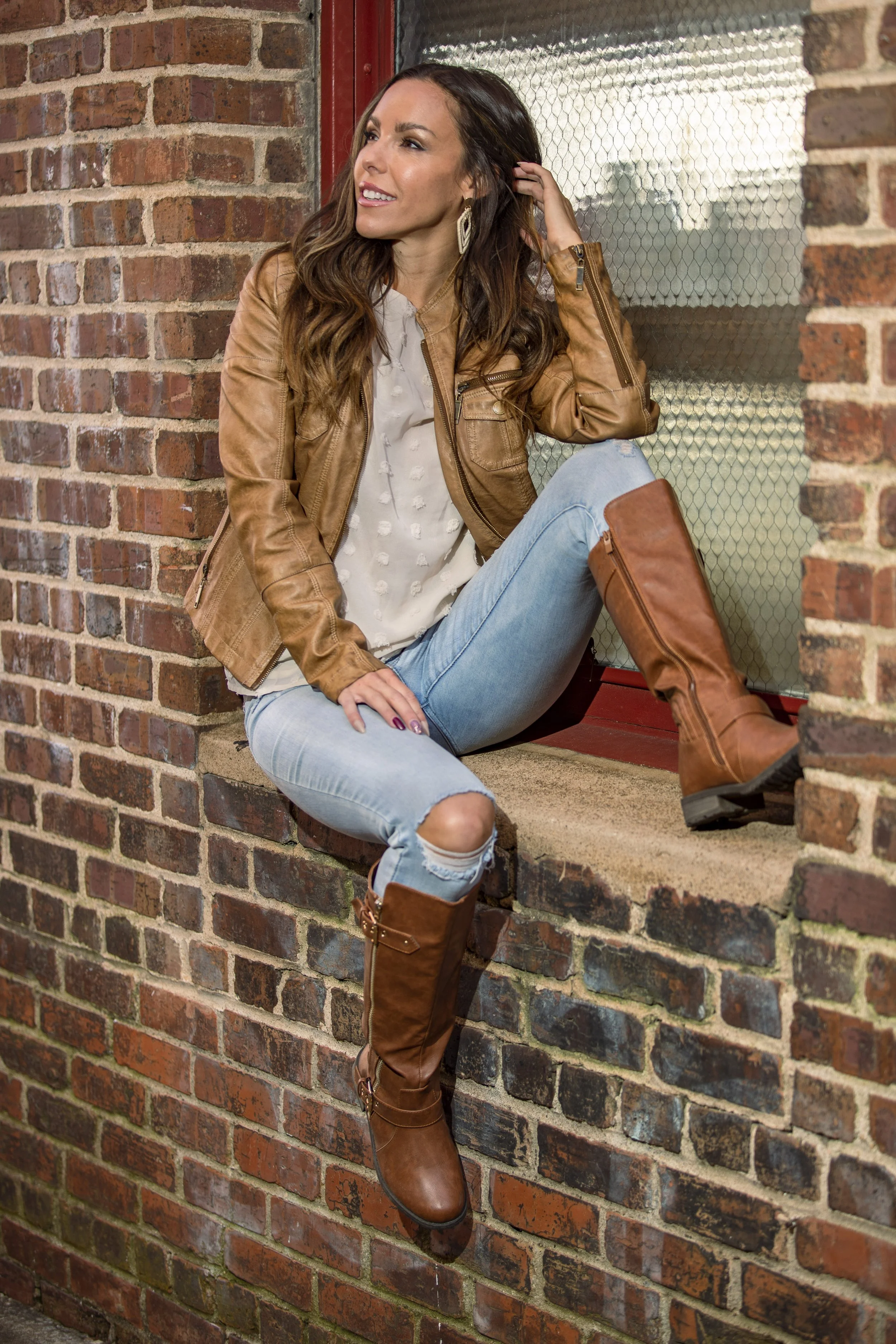 A woman sitting on a brick windowsill, wearing a brown leather jacket, light blue ripped jeans, and tall brown boots, looking to the side. BoldVision Studios