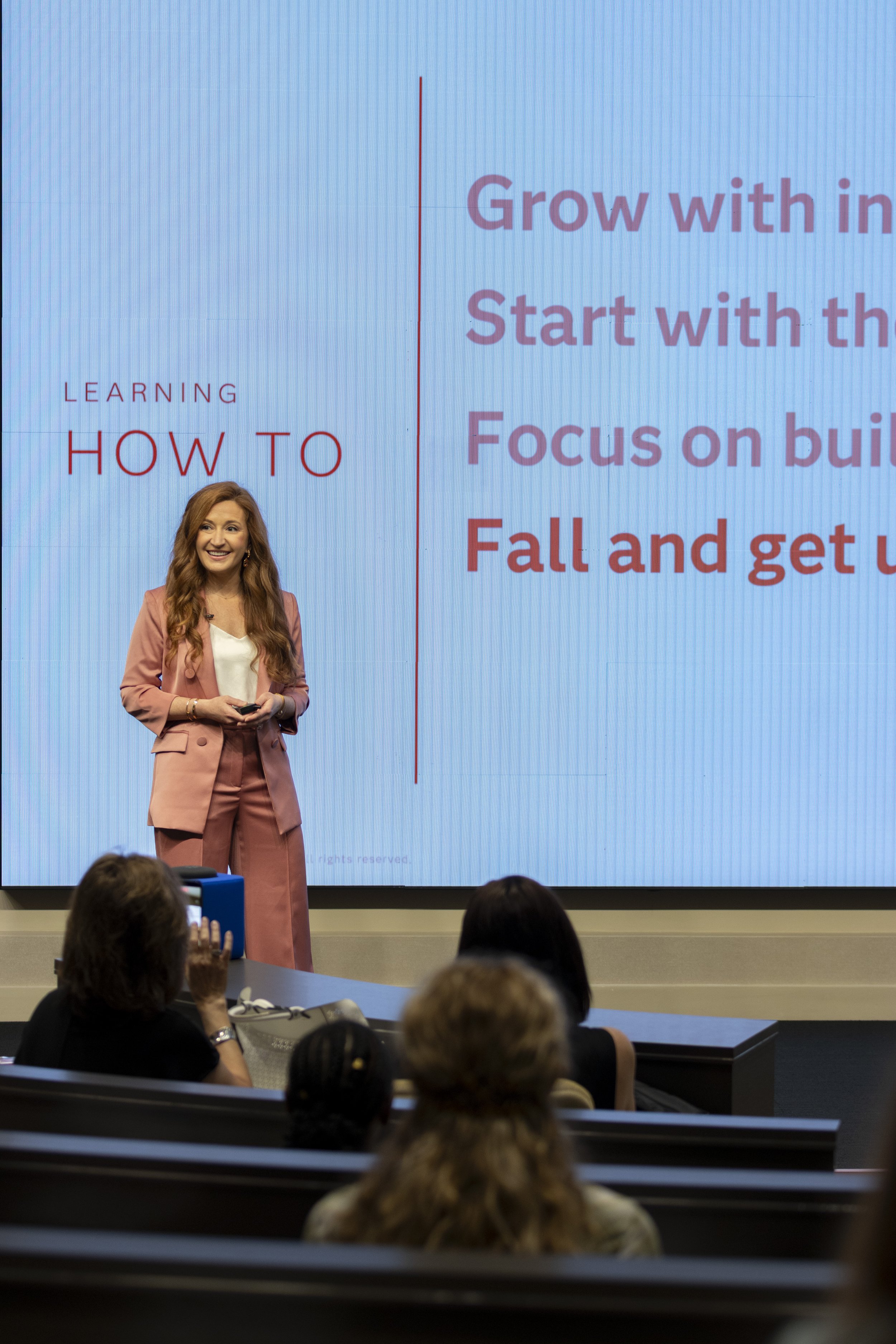 A woman with long red hair, wearing a pink blazer and matching pants, standing and speaking in front of an audience in a conference room. Behind her is a large screen displaying the words "LEARNING HOW TO" and additional text about growth, focusing, 