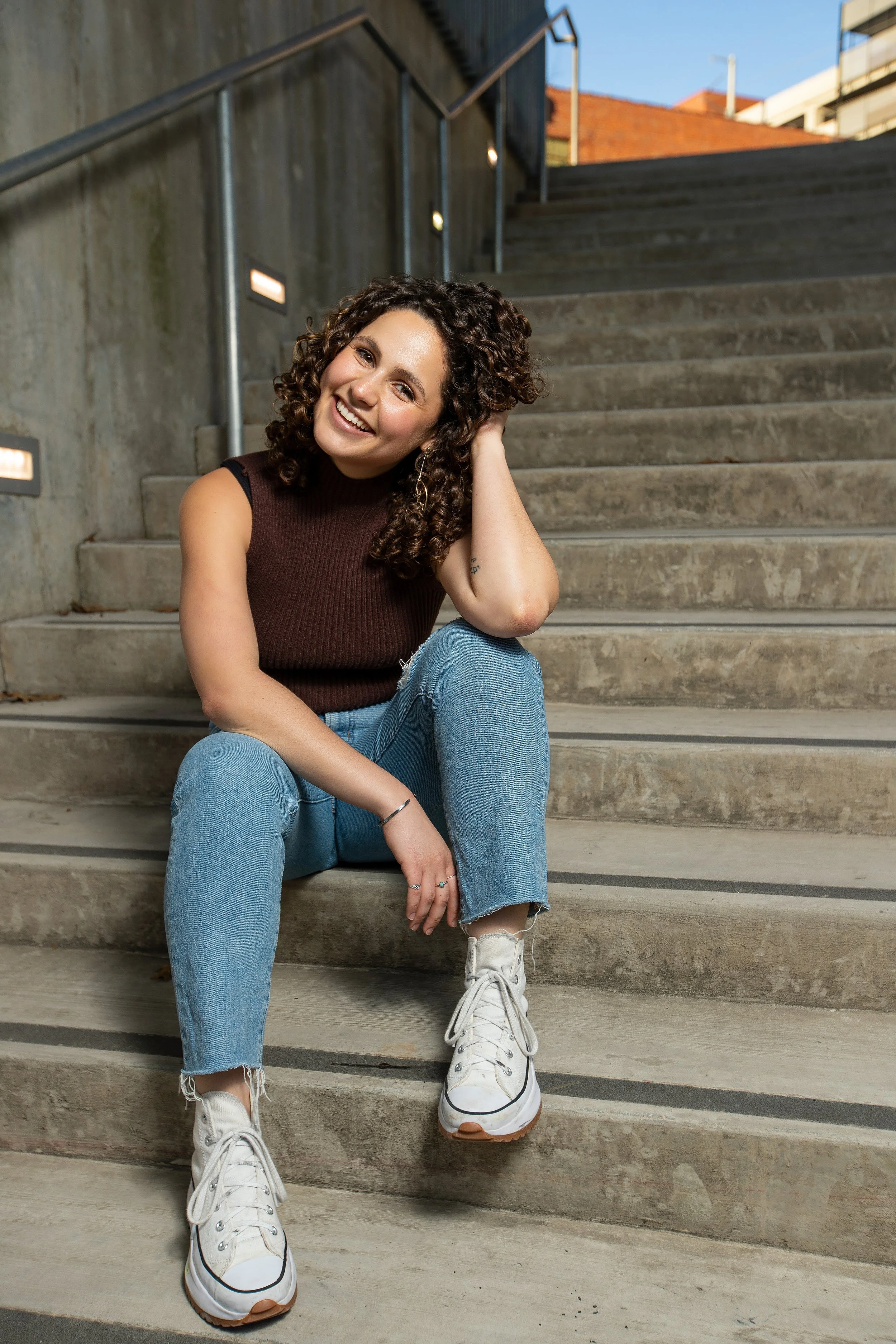 A young woman with curly hair sitting on concrete stairs outdoors, smiling, wearing a sleeveless brown top, blue jeans, and white sneakers, with a cityscape in the background. BoldVision Studios