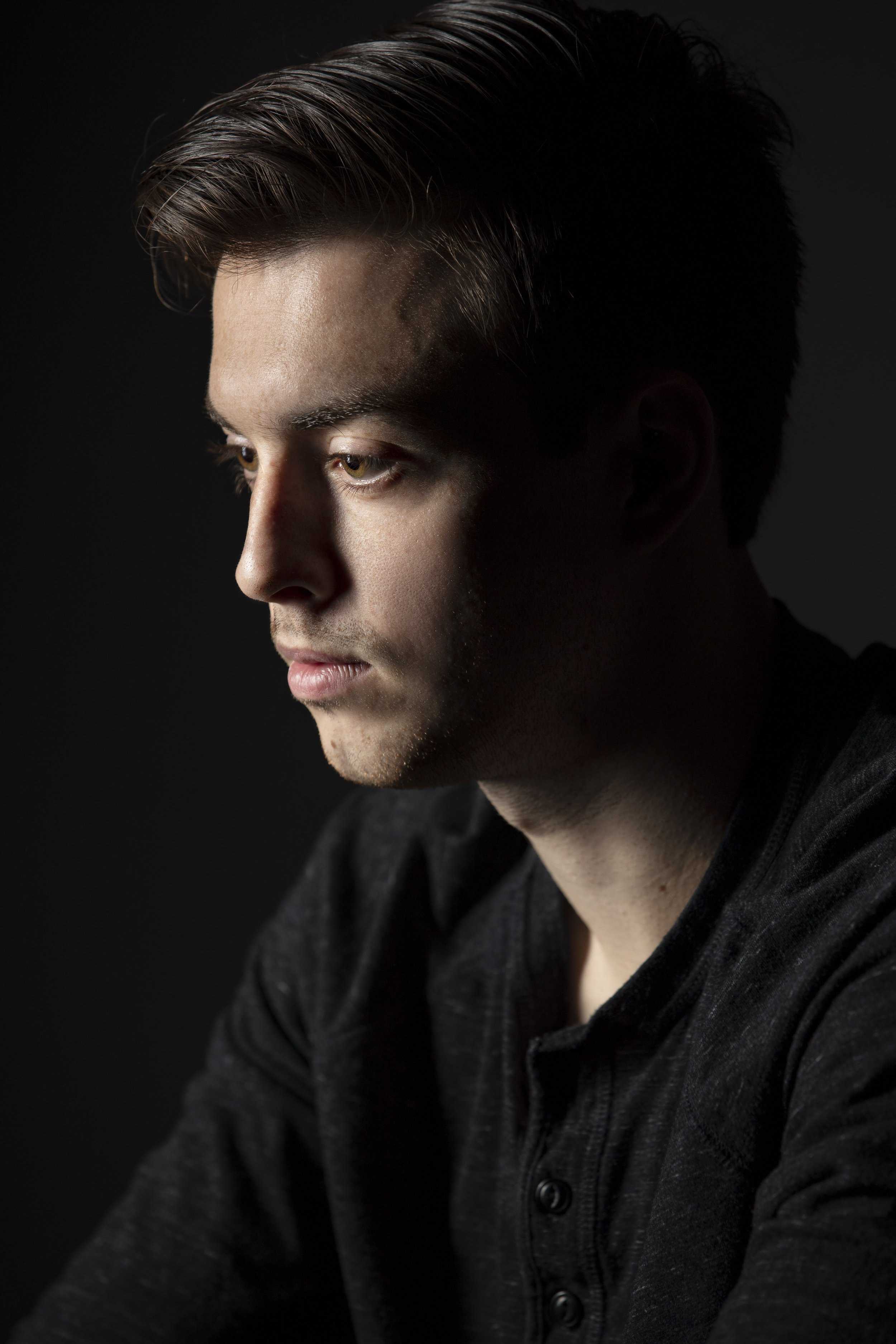 Profile of a young man with styled hair, looking down, wearing a dark shirt against a black background. BoldVision Studios
