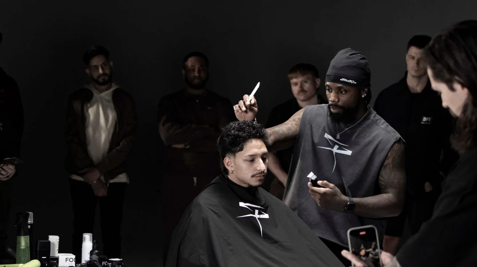 A man with tattoos, wearing a black beanie and sleeveless shirt, is giving a haircut to a young man with curly hair, who is sitting with a black cape. Several people are watching in the background, and there are various grooming products on a table i