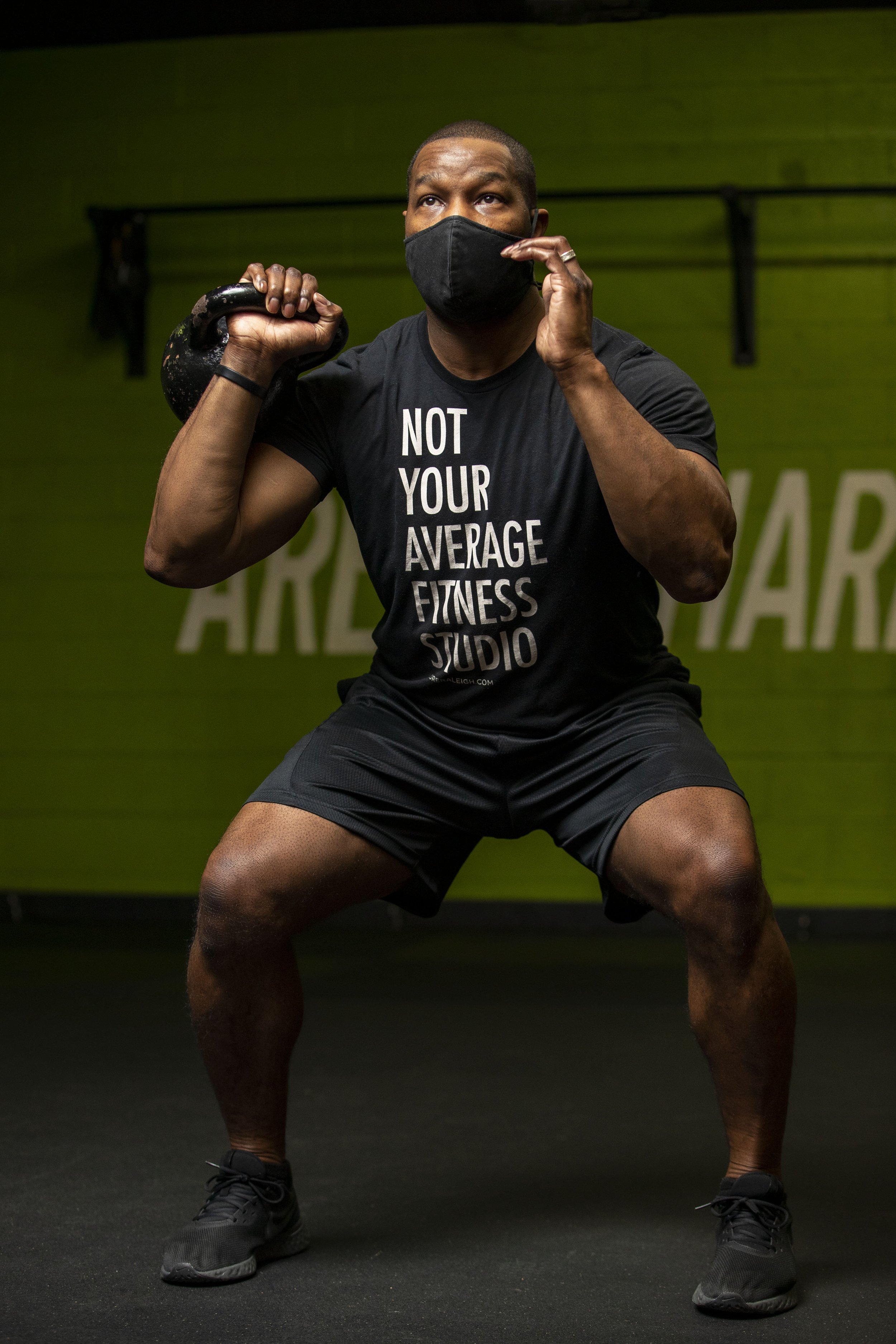 A man in black workout attire and a black face mask is holding a kettlebell on his shoulder during exercise, standing in a gym with green walls. BoldVision Studios