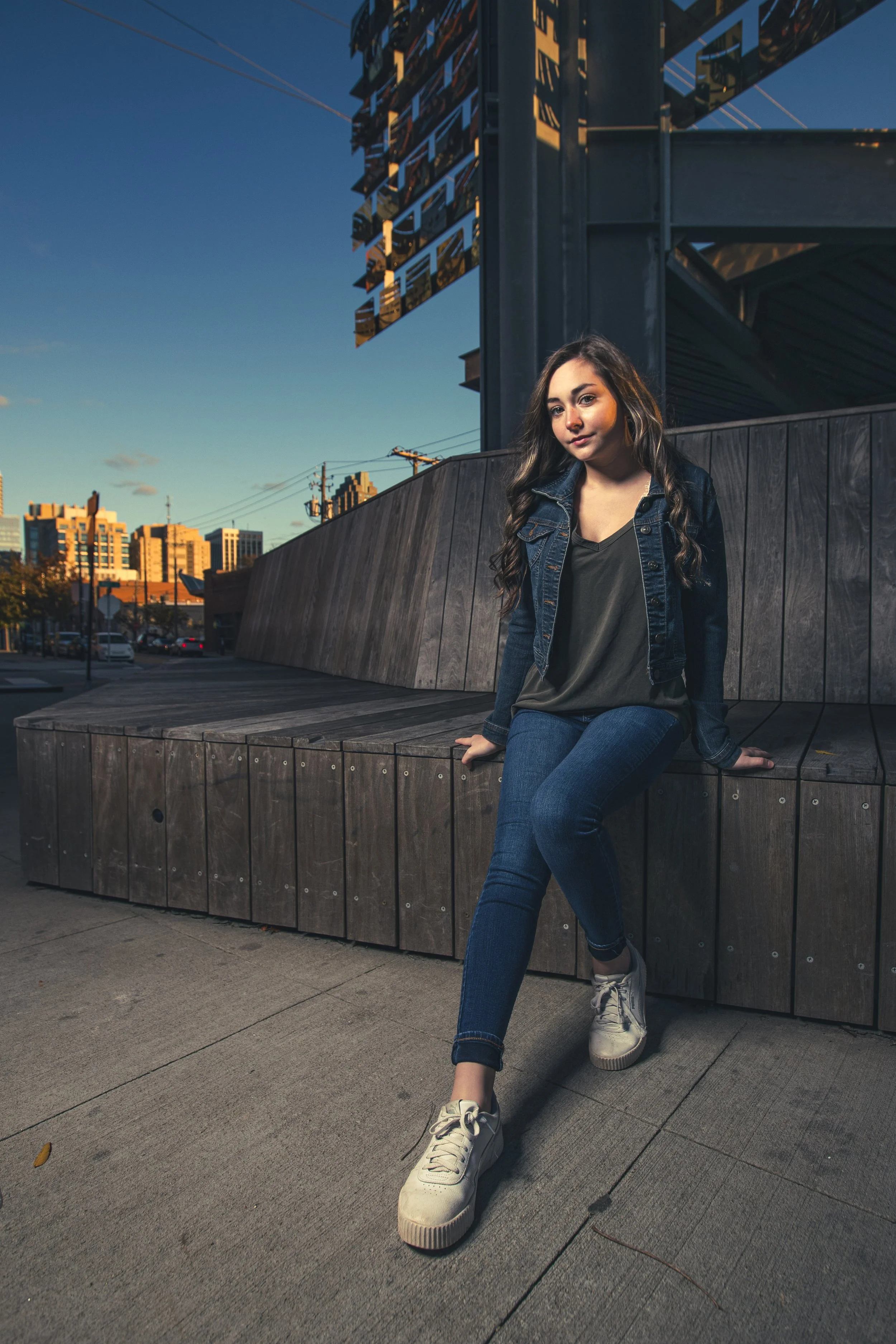 Young woman sitting on a wooden bench outdoors during sunset with a cityscape in the background. BoldVision Studios