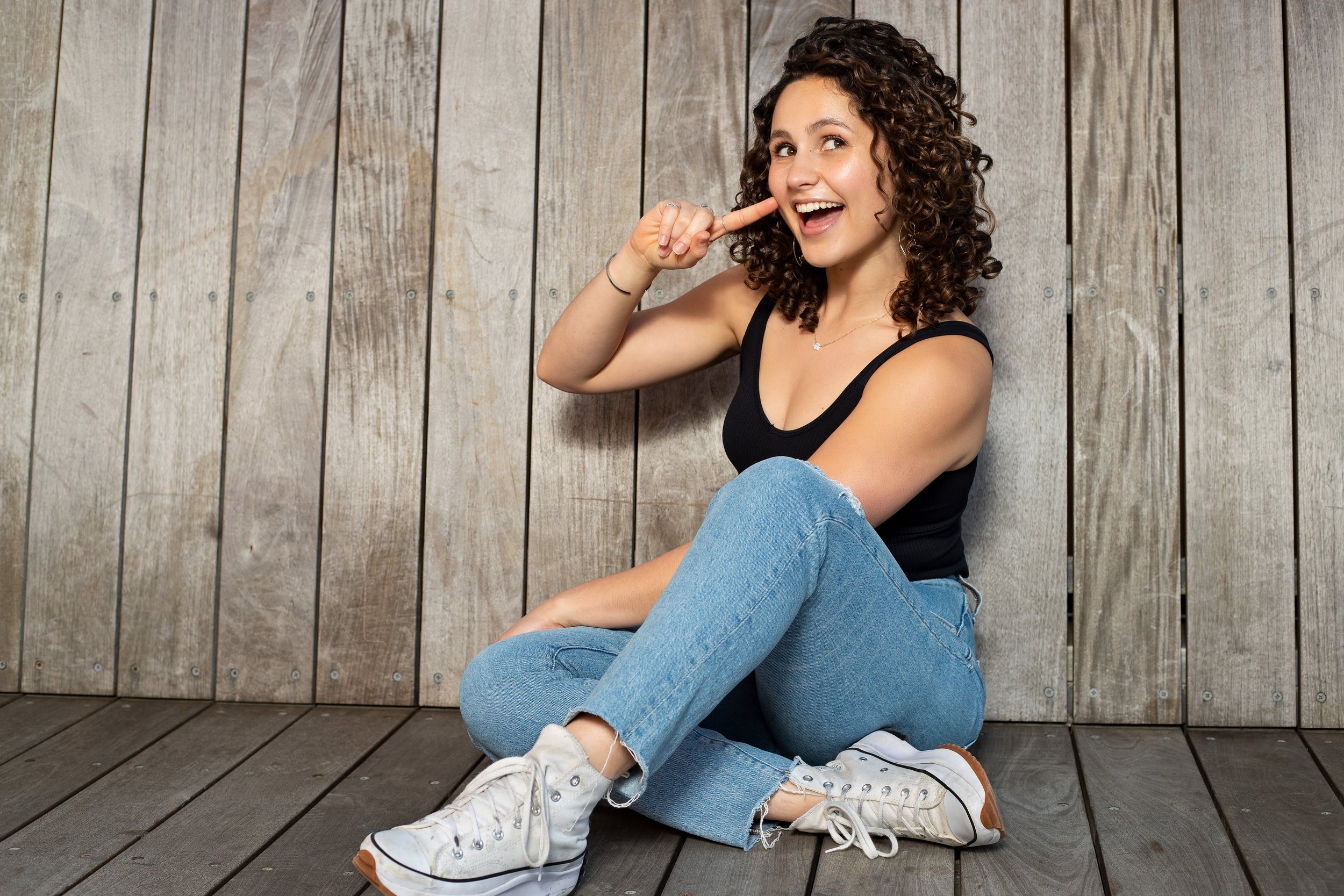 A young woman sitting on a wooden floor, posing with a playful smile, wearing a black tank top, ripped blue jeans, and white sneakers, with curly brown hair, against a wooden paneled wall. BoldVision Studios
