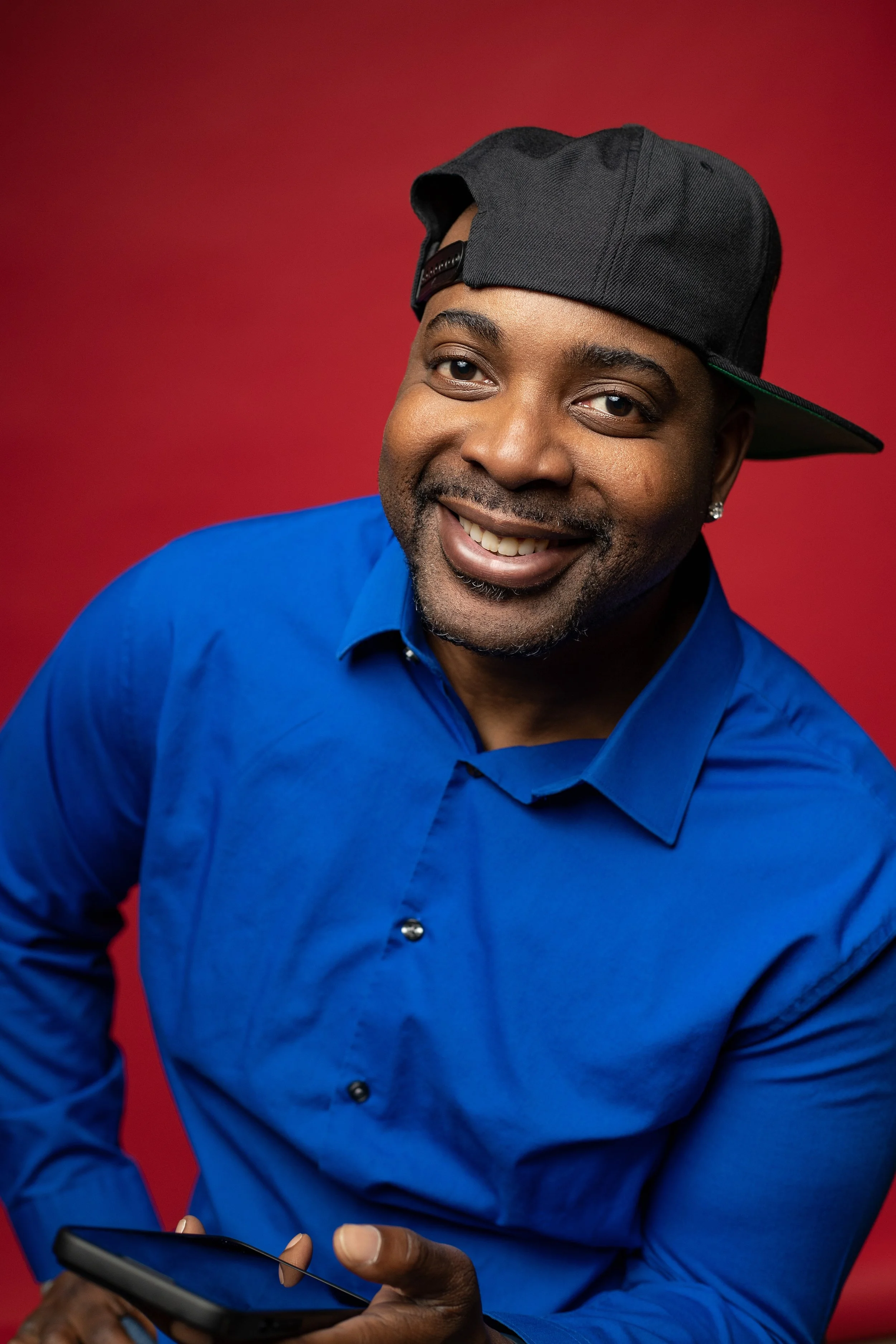A smiling man wearing a blue dress shirt and a black cap, holding a smartphone, against a red background. BoldVision Studios