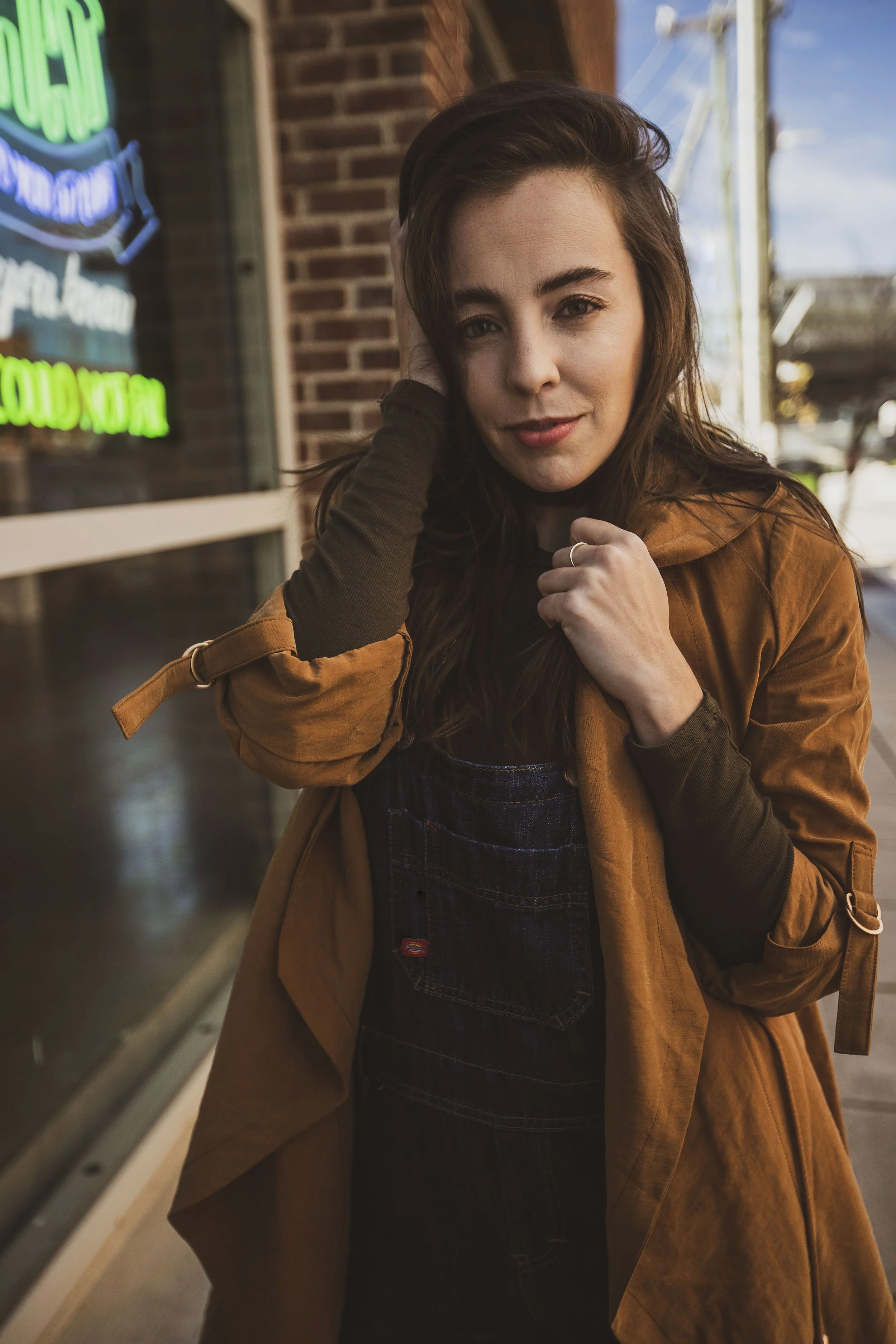 A woman with brown hair, wearing a brown jacket and black overalls, standing outside near a brick building with neon signs. She is touching her hair with her left hand and looking at the camera. BoldVision Studios