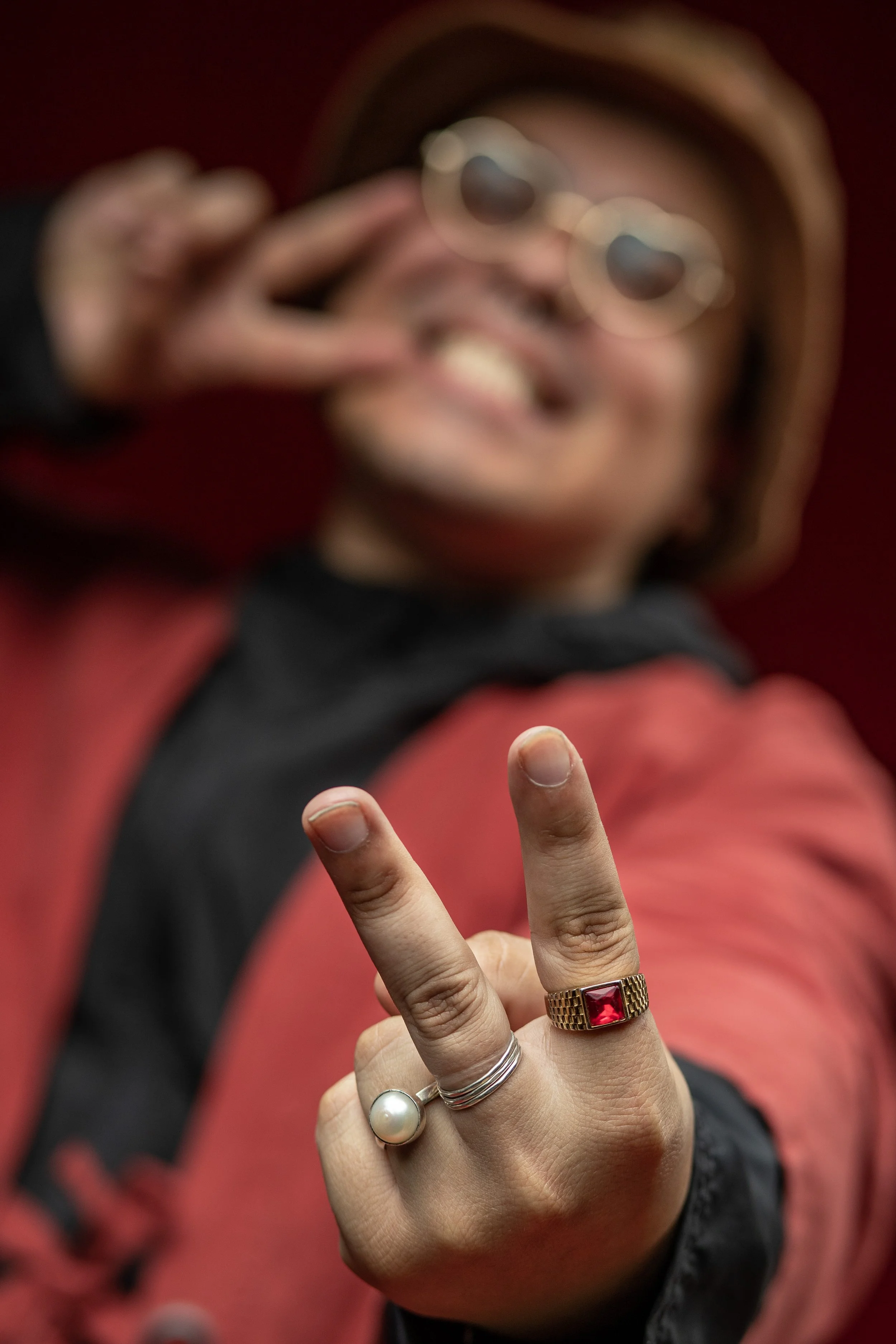 Blurred image of a smiling person wearing sunglasses, a hat, and a red jacket, making a peace sign with their hand which has a large pearl ring and a red gemstone ring. BoldVision Studios