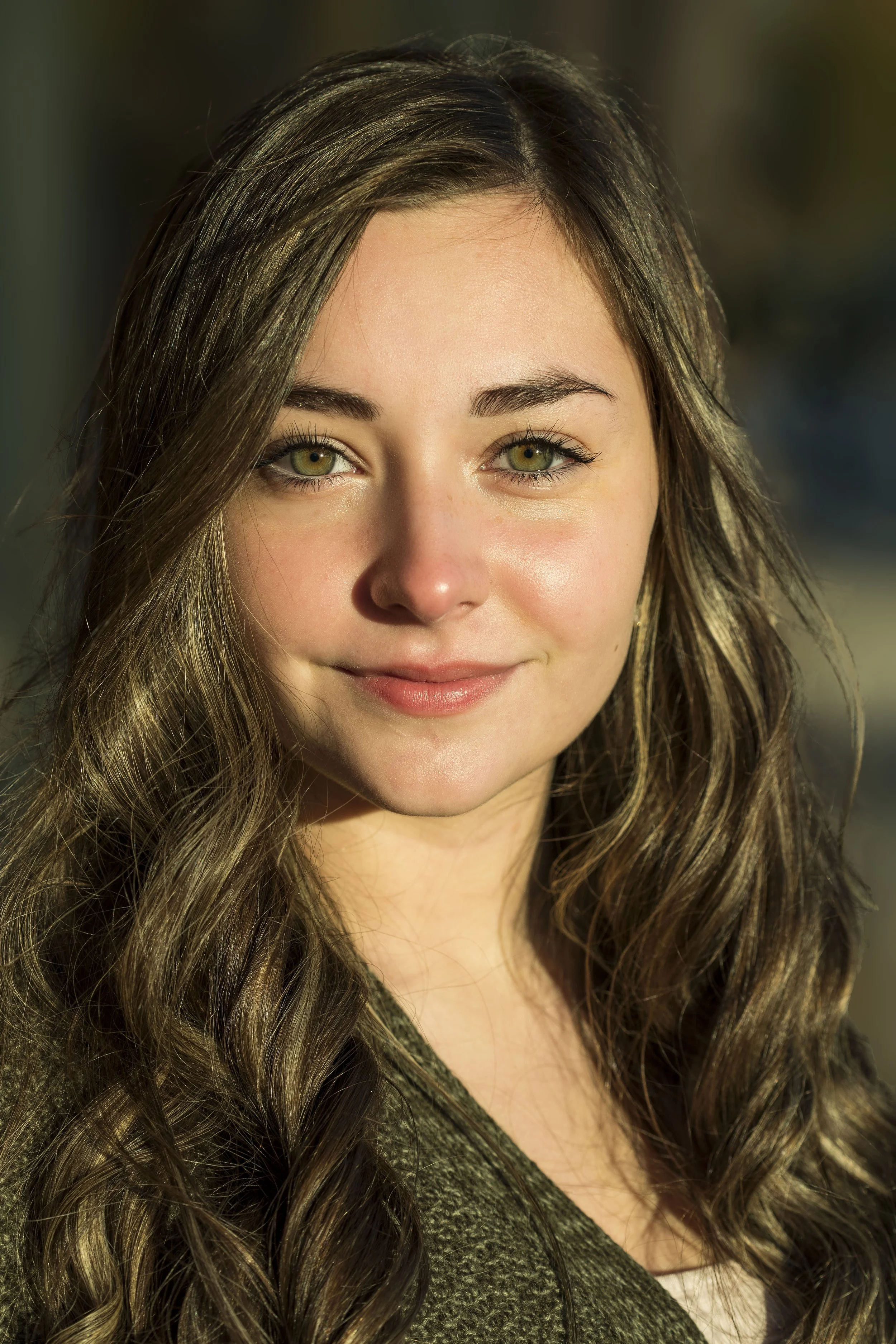 Close-up portrait of a young woman with long, wavy brown hair and green eyes, smiling softly outdoors with sunlight on her face. BoldVision Studios