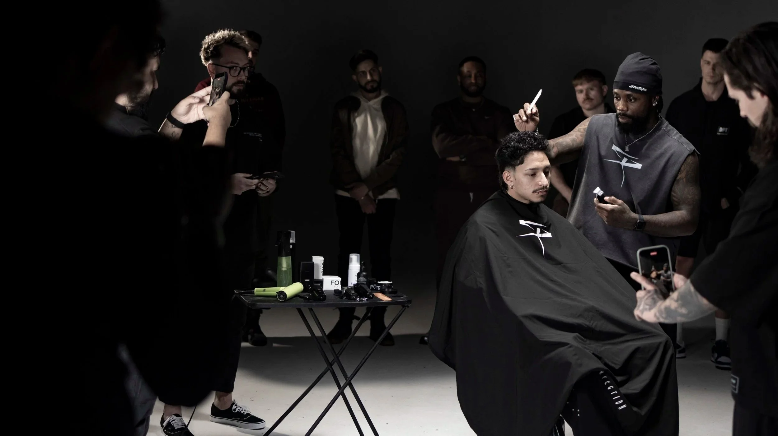 A man is getting a haircut or haircut consultation in a dark room while multiple people around him are taking photos or videos with their smartphones. A table with hair styling tools and products is in front of him.