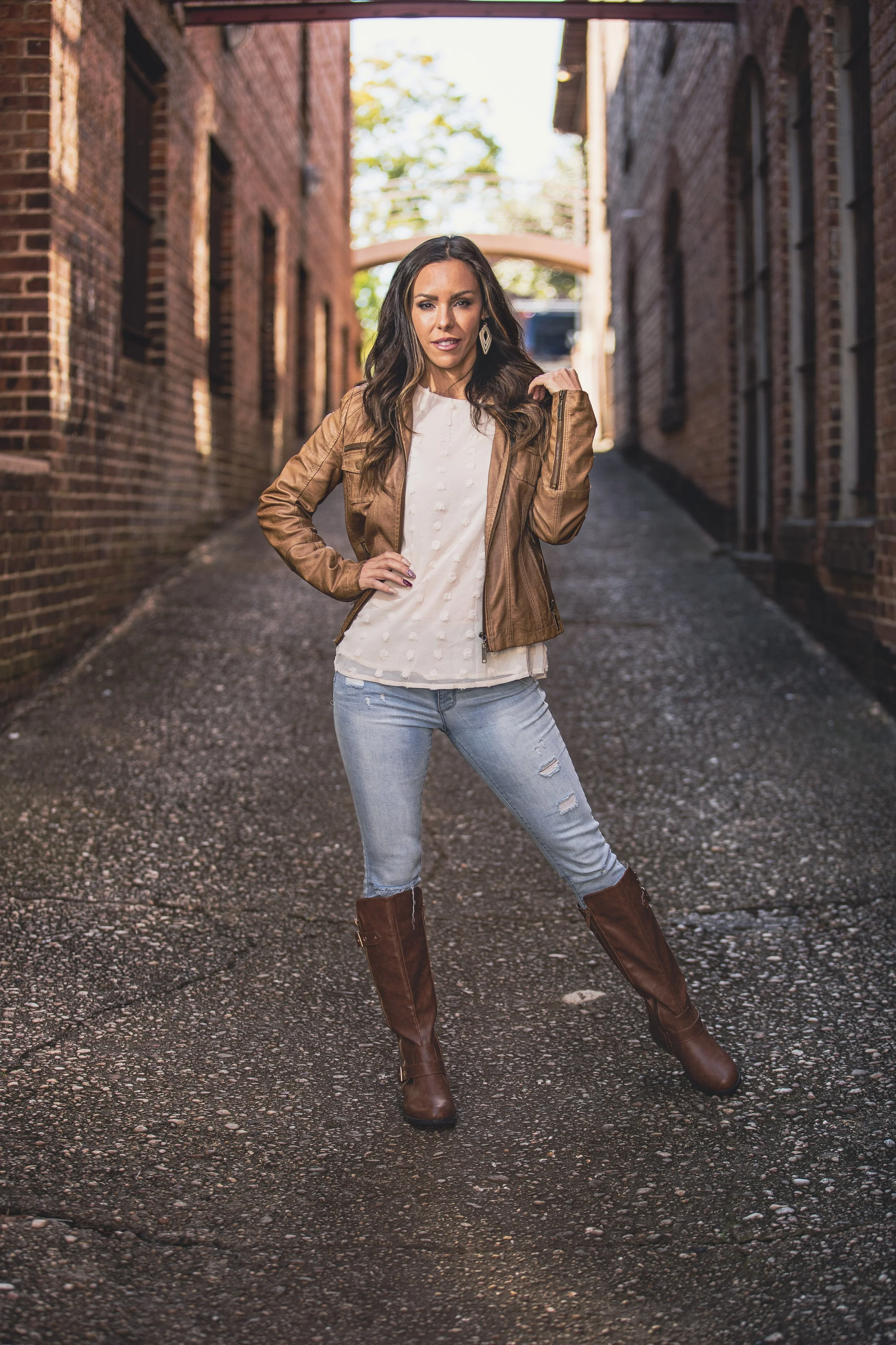 A woman stands confidently in a narrow alleyway with brick walls on each side, wearing a brown jacket, white top, distressed jeans, and tall brown boots. BoldVision Studios