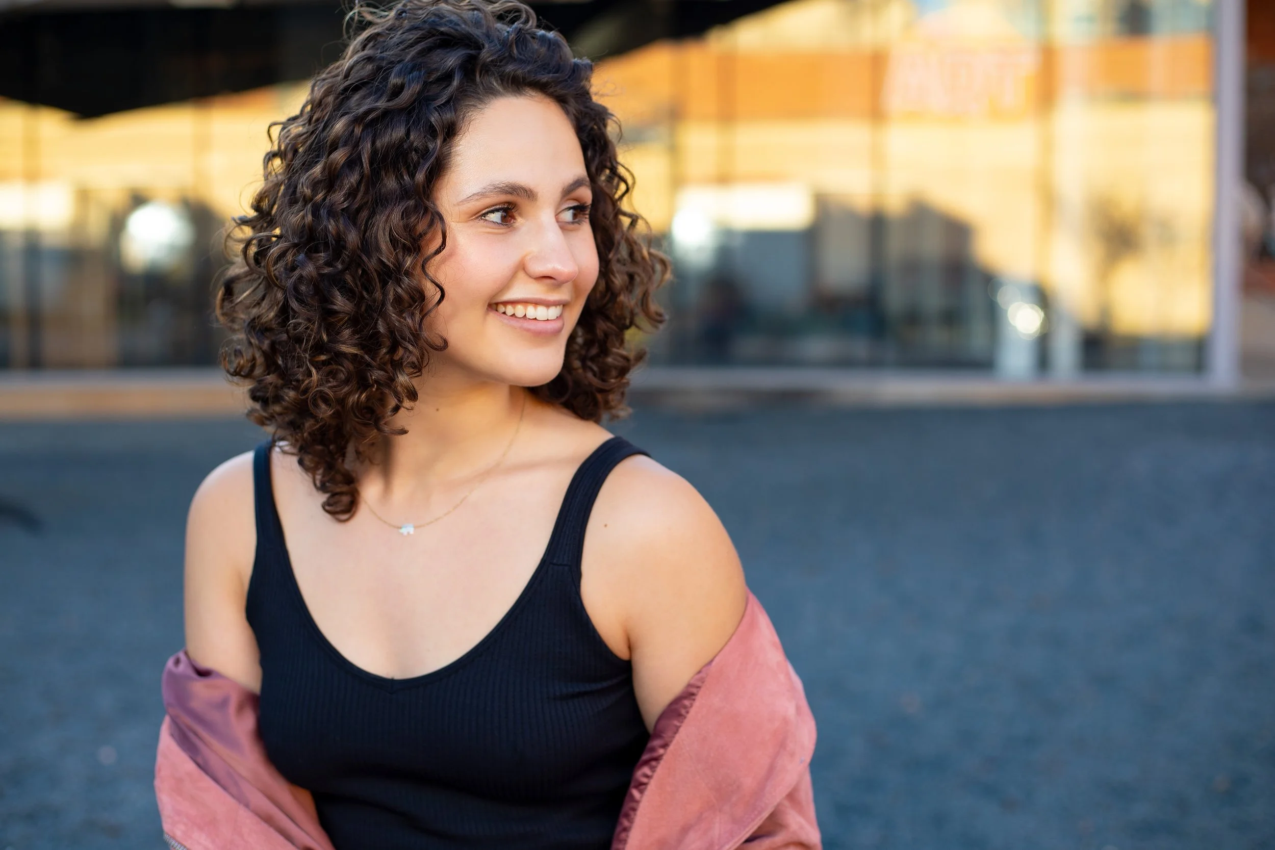 Young woman with curly hair smiling outdoors, wearing a black tank top and a pink jacket draped over her arms, standing in front of a building with reflective windows. BoldVision Studios