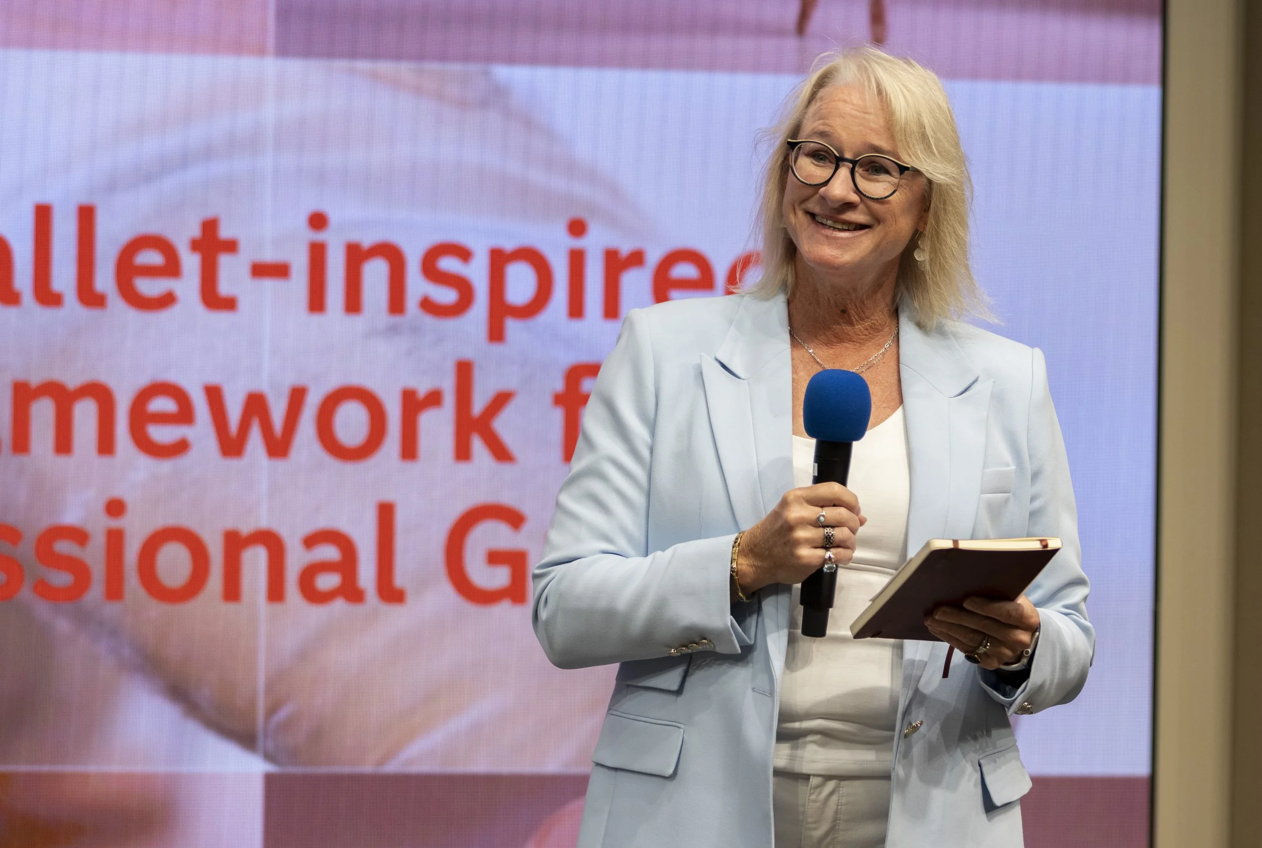 A woman with blonde hair and glasses, wearing a light blue blazer, holding a microphone and a tablet, standing in front of a large screen with red text.