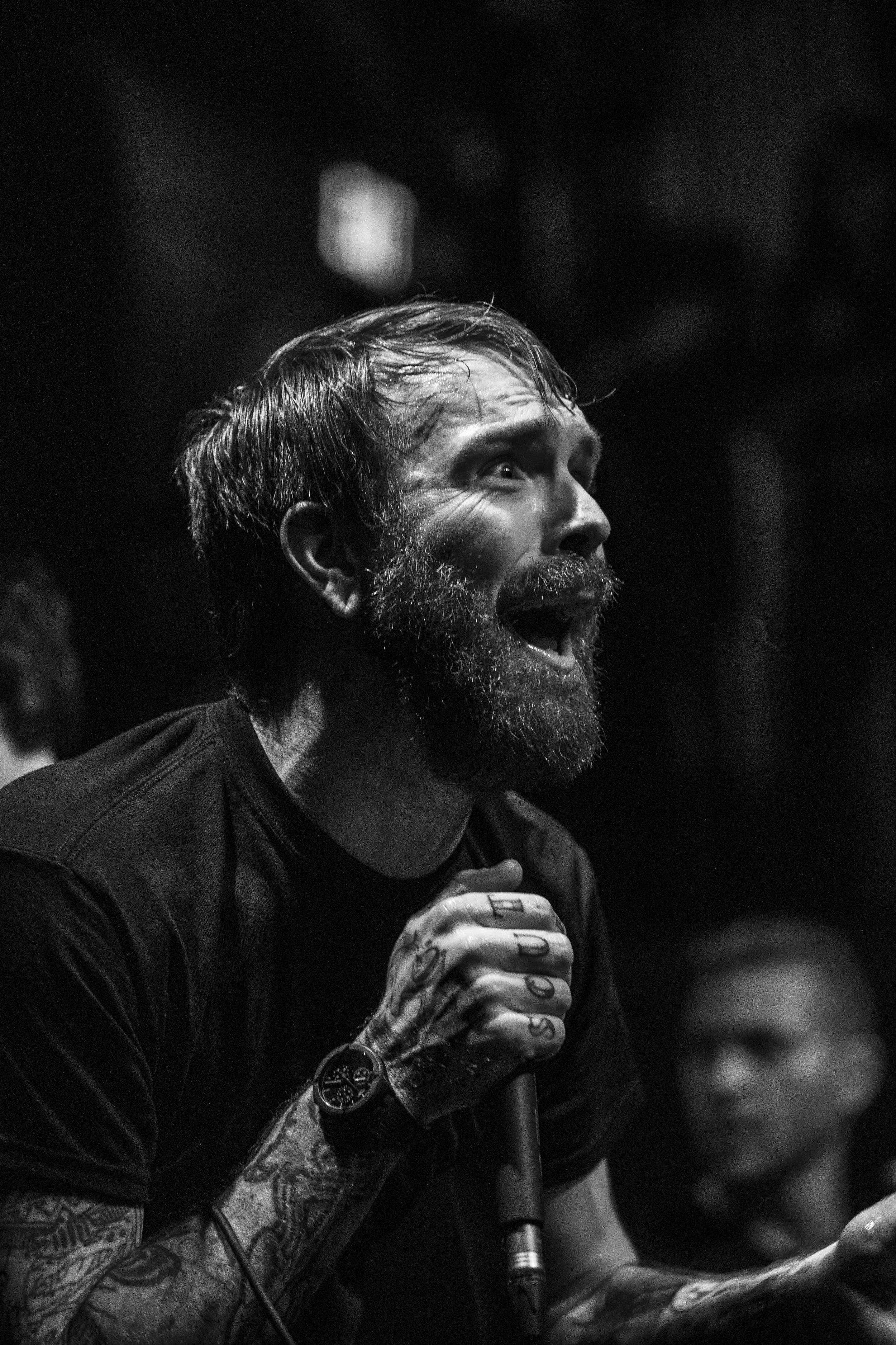 A man with a beard and tattoos singing passionately into a microphone in a dark setting, with a woman blurred in the background. BoldVision Studios