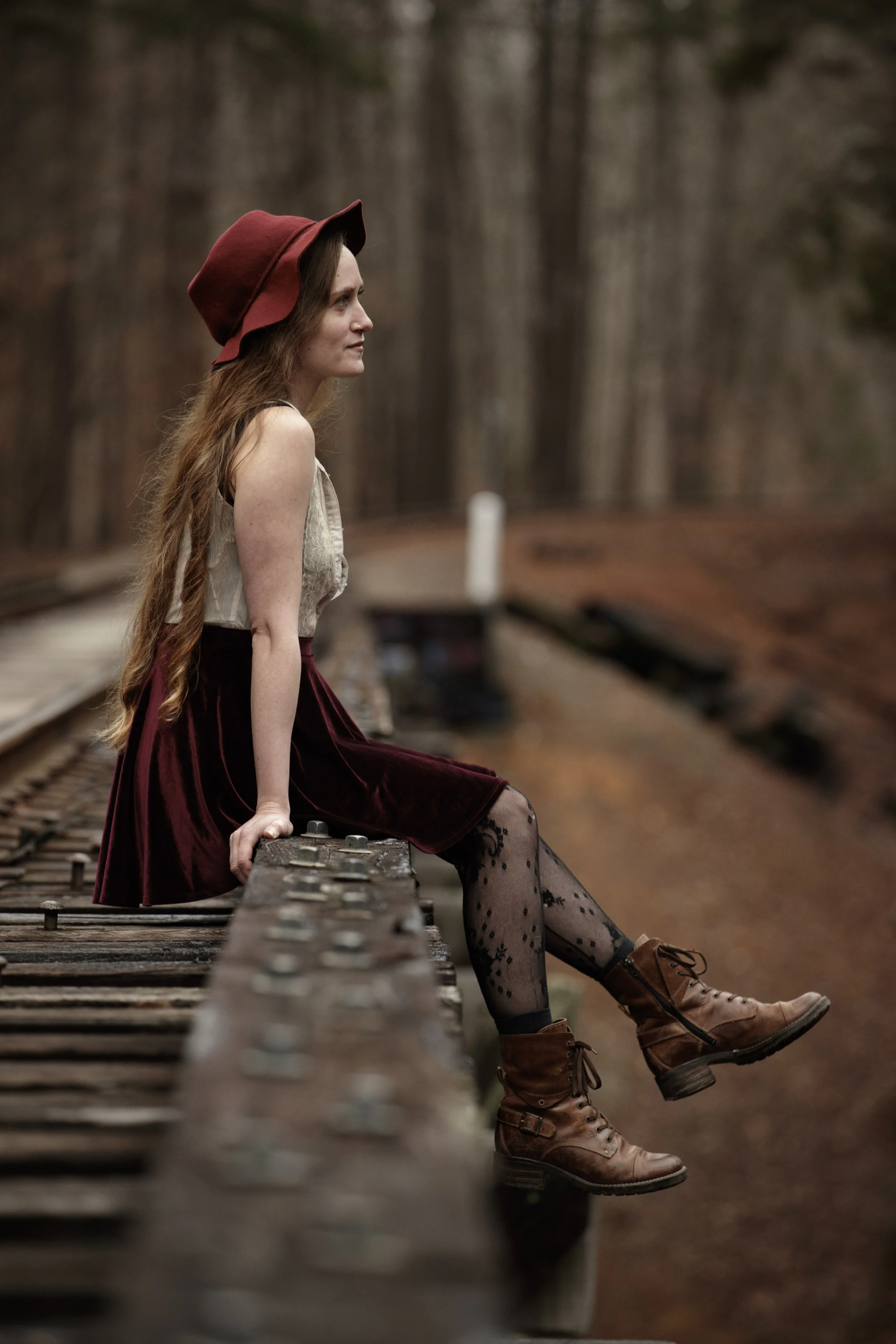 A woman with long hair, wearing a red hat, sleeveless top, a red velvet skirt, patterned tights, and brown boots, sitting on the edge of a railroad track, looking into the distance in a wooded area. BoldVision Studios