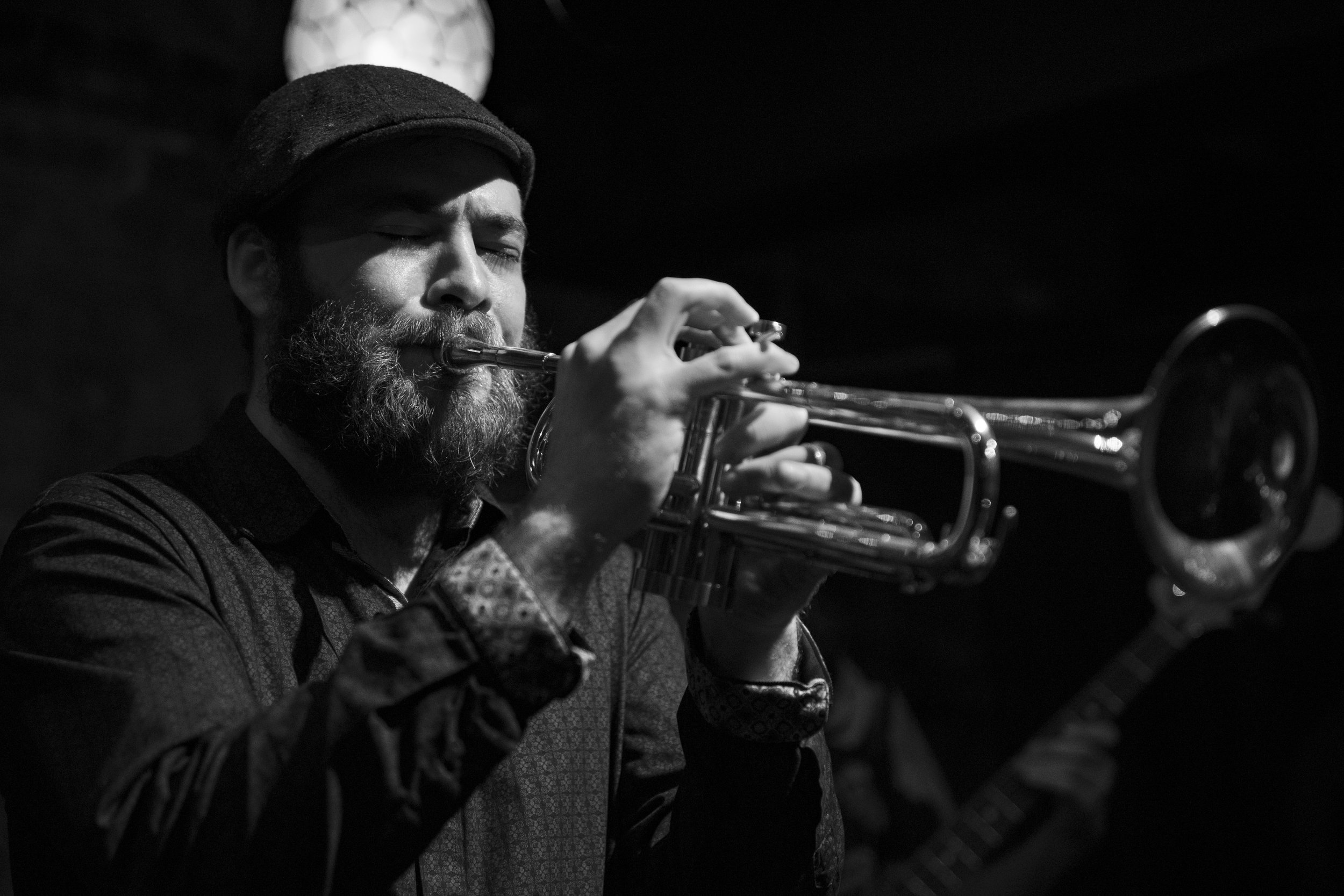A man with a beard and a hat playing a trumpet in a low-light setting. BoldVision Studios