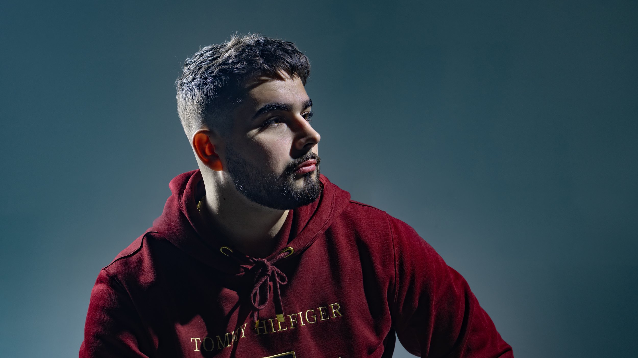 A young man with a beard and short dark hair, wearing a red Tommy Hilfiger hoodie, looking to the right against a plain background. BoldVision Studios