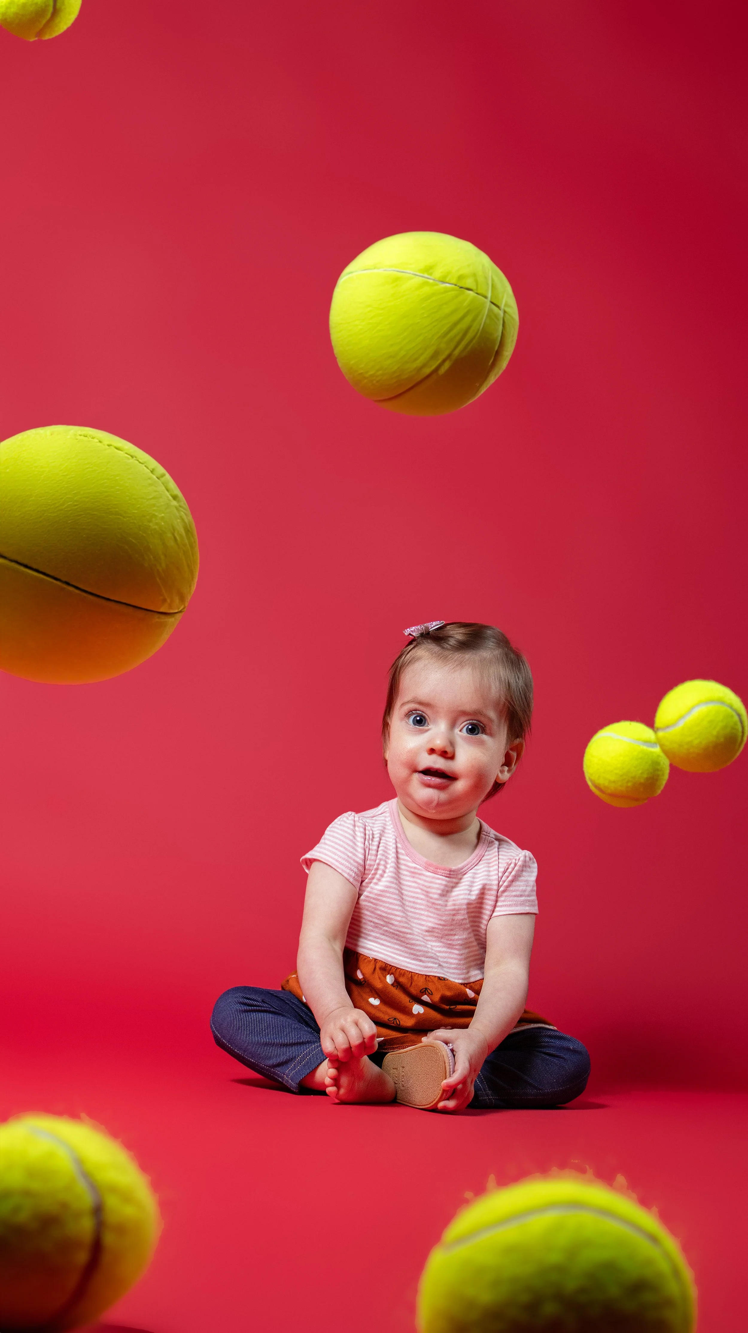 A young girl sitting on a red background surrounded by yellow tennis balls, with additional tennis balls floating in the air.