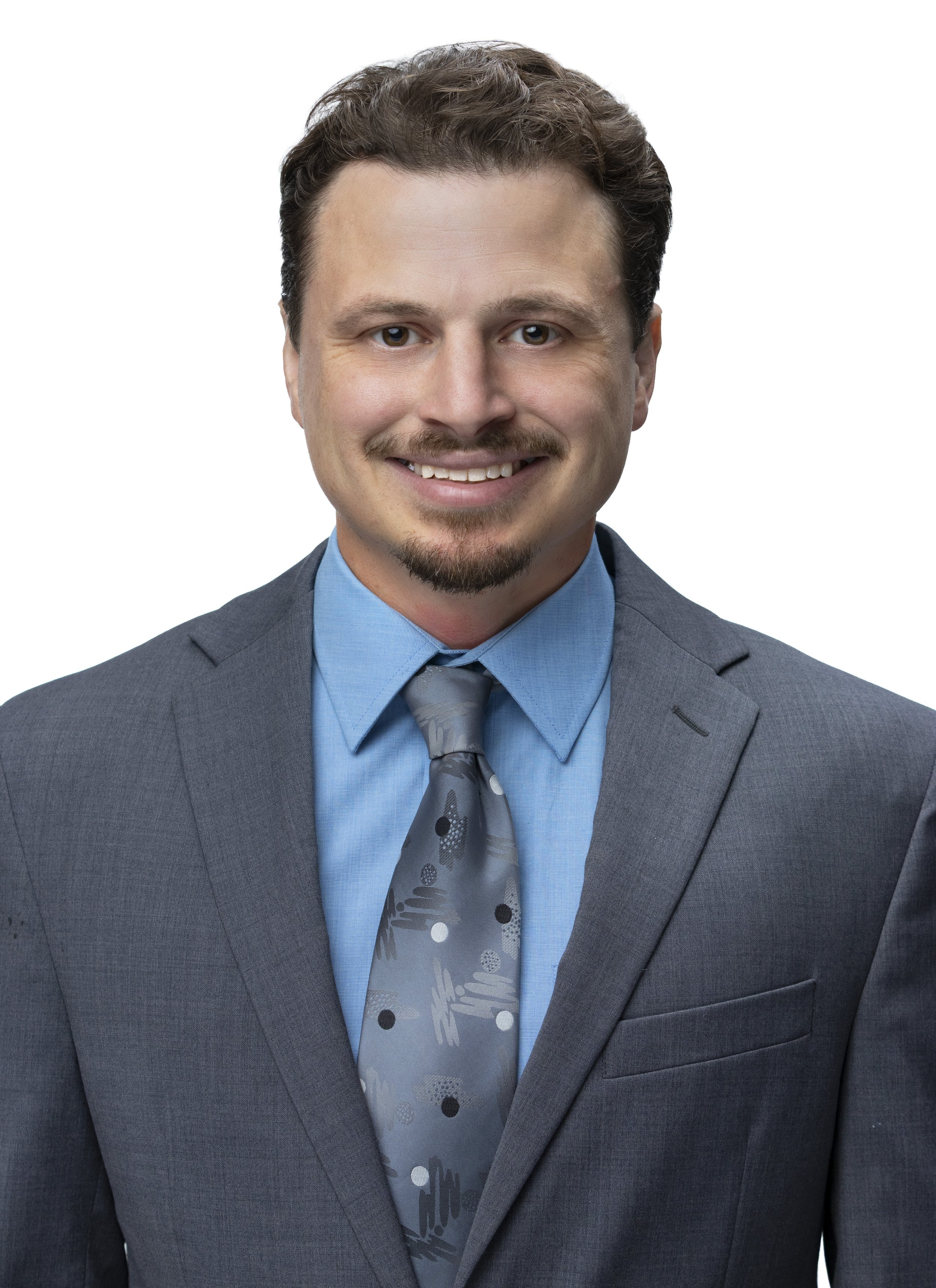 A man in a gray suit, blue shirt, and patterned gray tie smiling, with brown hair and a goatee, against a white background.