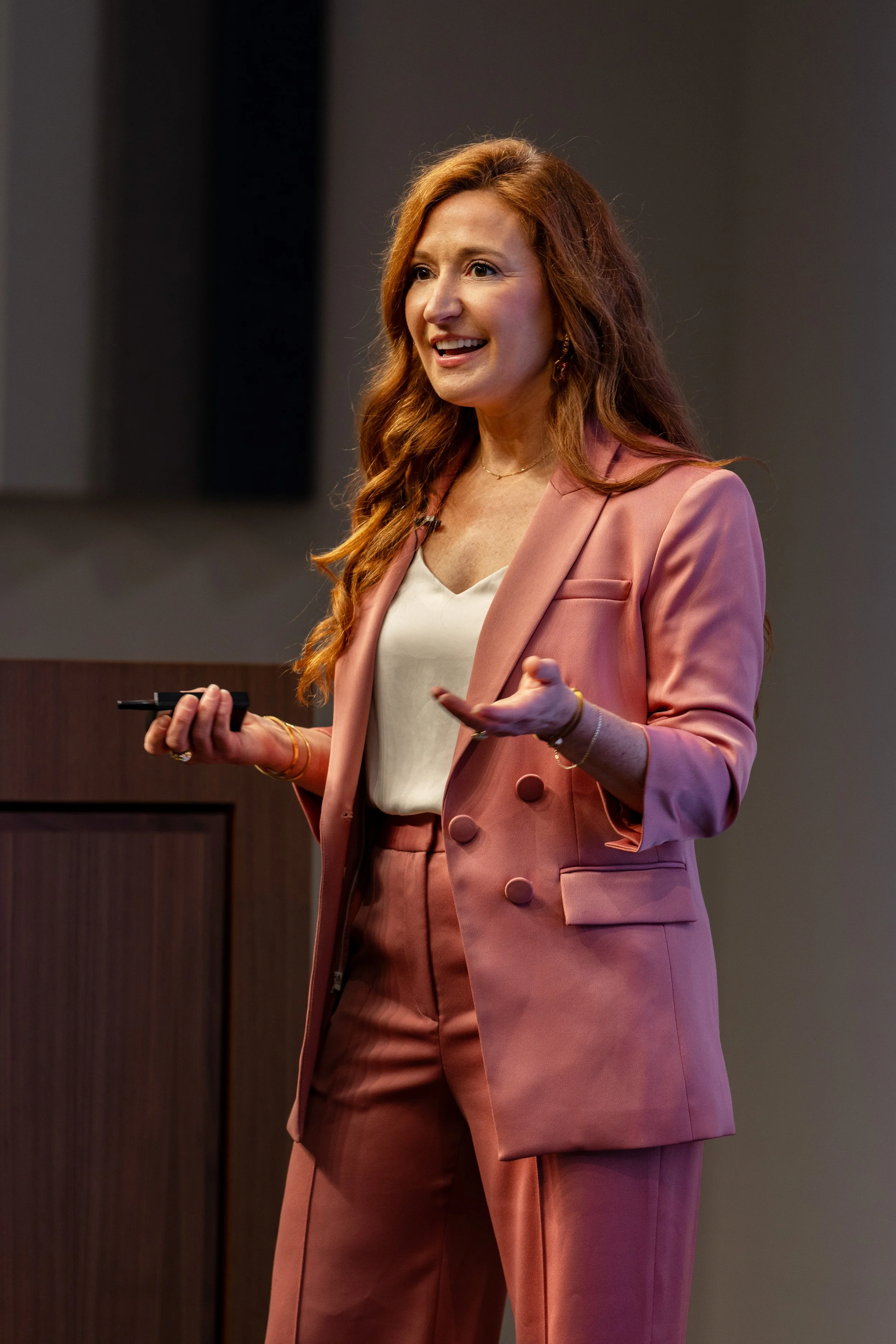 A woman with long reddish hair, wearing a pink blazer and pink pants, giving a presentation in a professional setting.
