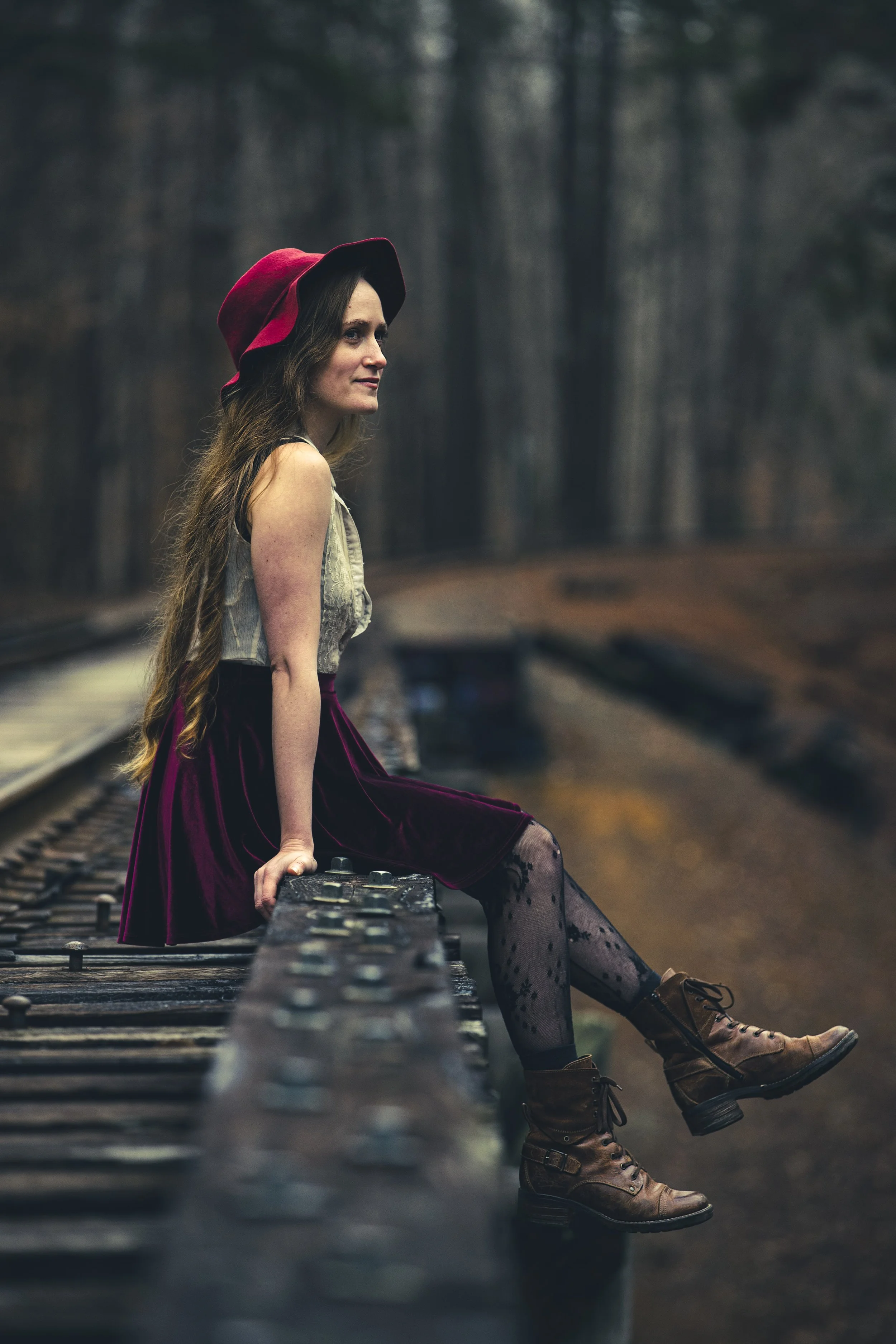 A woman with long hair, wearing a red hat, gray top, and maroon skirt, sitting on a railway track in a forest, with her feet resting on a rail. BoldVision Studios