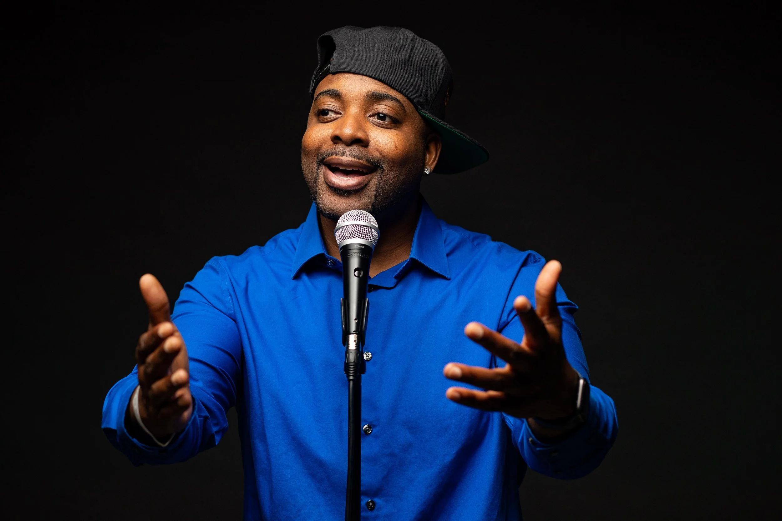 A man wearing a blue shirt and a black cap is standing in front of a black background, speaking into a microphone, with a smile and gesturing with his hands. BoldVision Studios