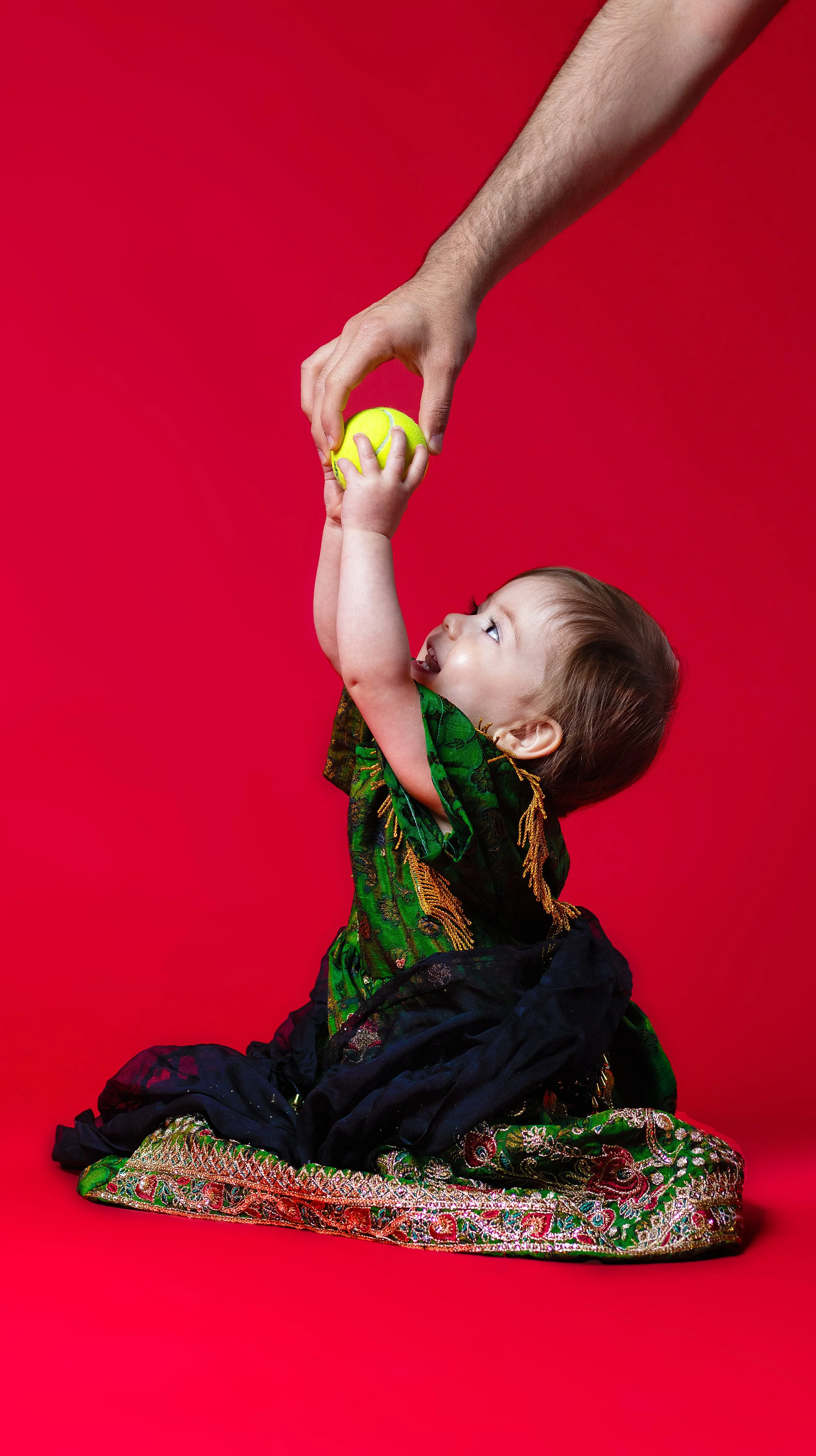 A young child in traditional colorful clothing reaches up to catch a yellow tennis ball being handed to them by an adult, against a bright red background. BoldVision Studios